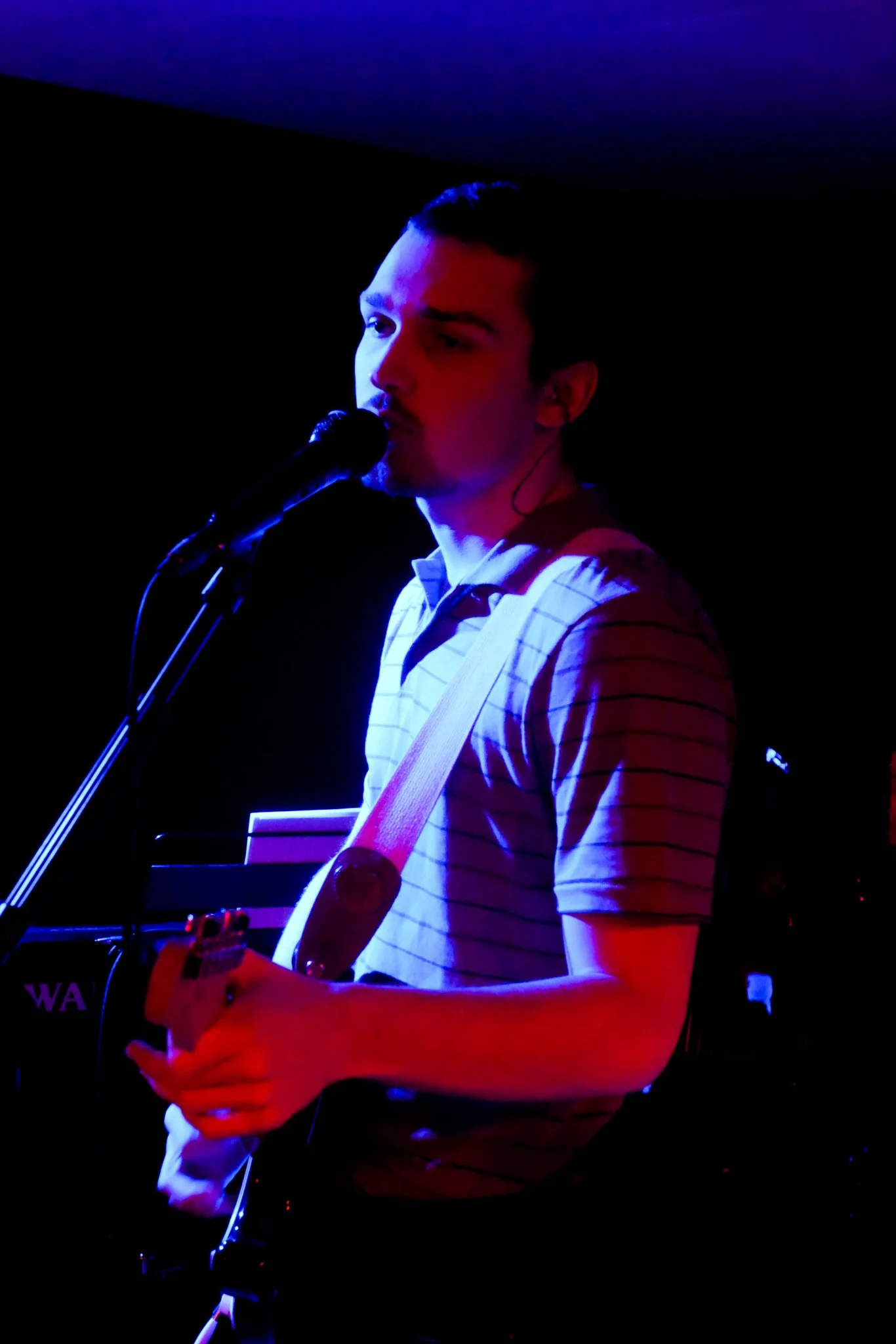 A young man with short hair, wearing a striped shirt, is performing live on stage with a guitar, singing into a microphone. The stage is lit with deep blue and purple lighting, creating a vibrant atmosphere.