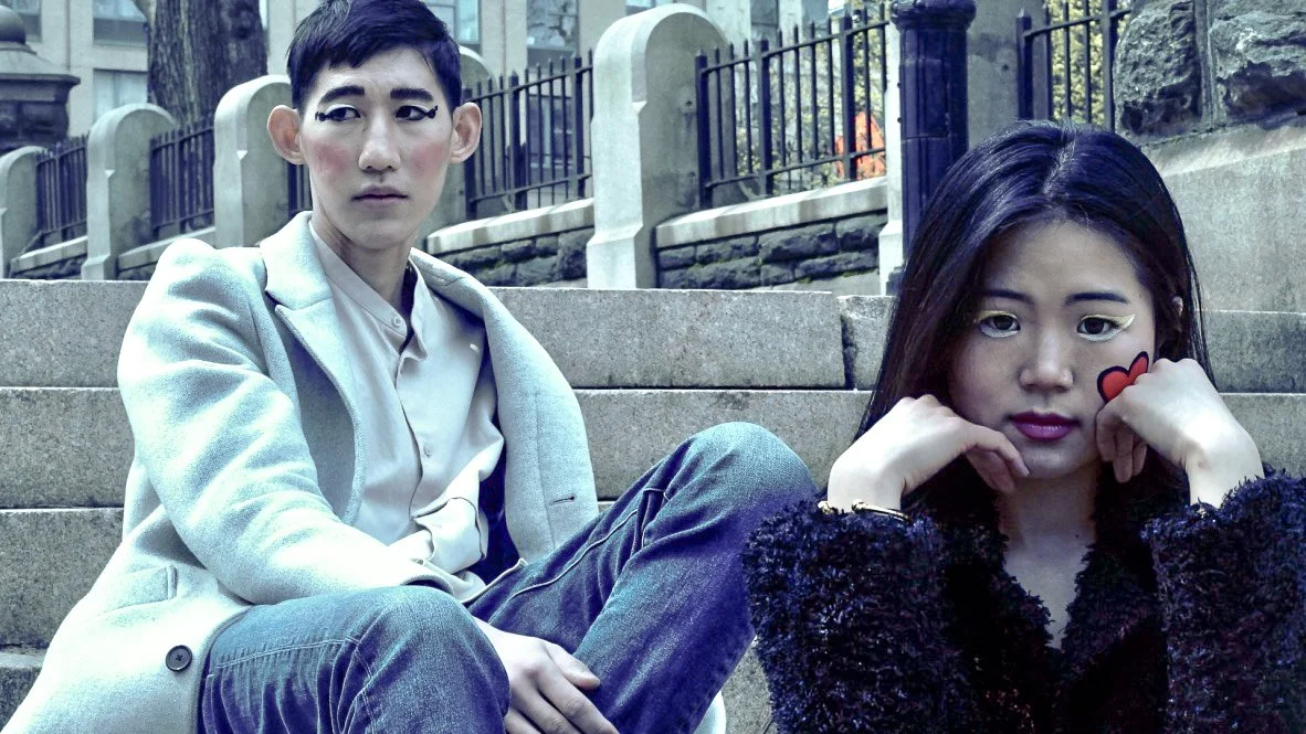 A young man and woman sitting on stone steps outdoors, with a fence and trees in the background. The man has short dark hair, makeup, and is wearing a light gray blazer and jeans. The woman has long dark hair, makeup with winged eyeliner, and has a s