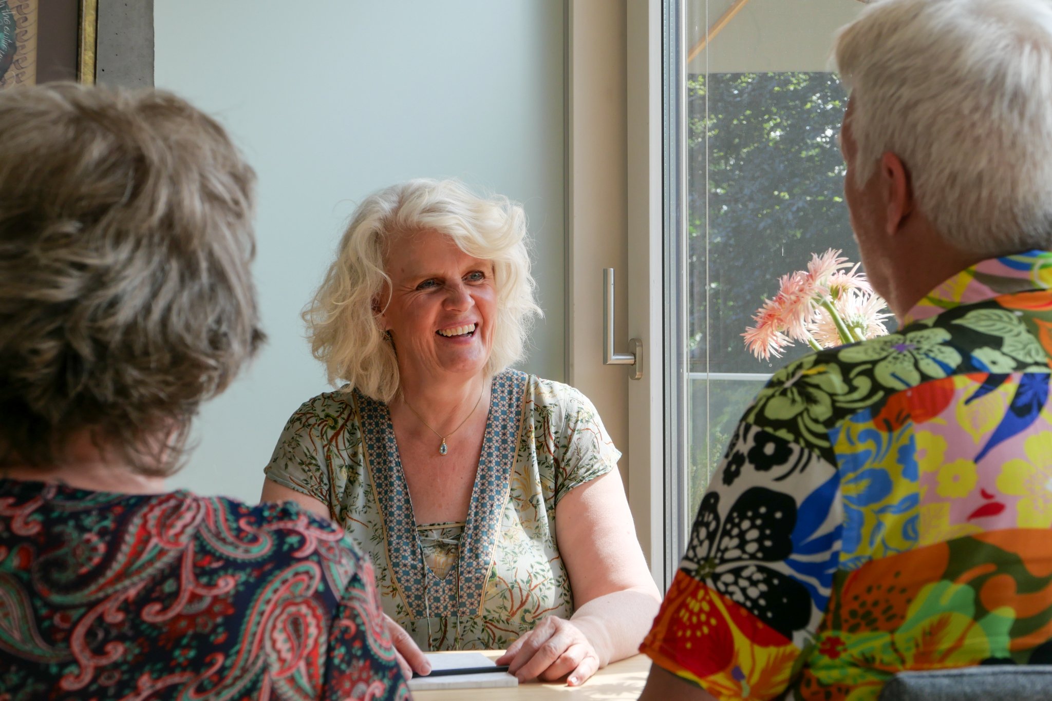 A woman with blonde curly hair smiling and engaging in conversation with a group of people in colorful clothing, sitting at a table near a window with a view of trees outside.