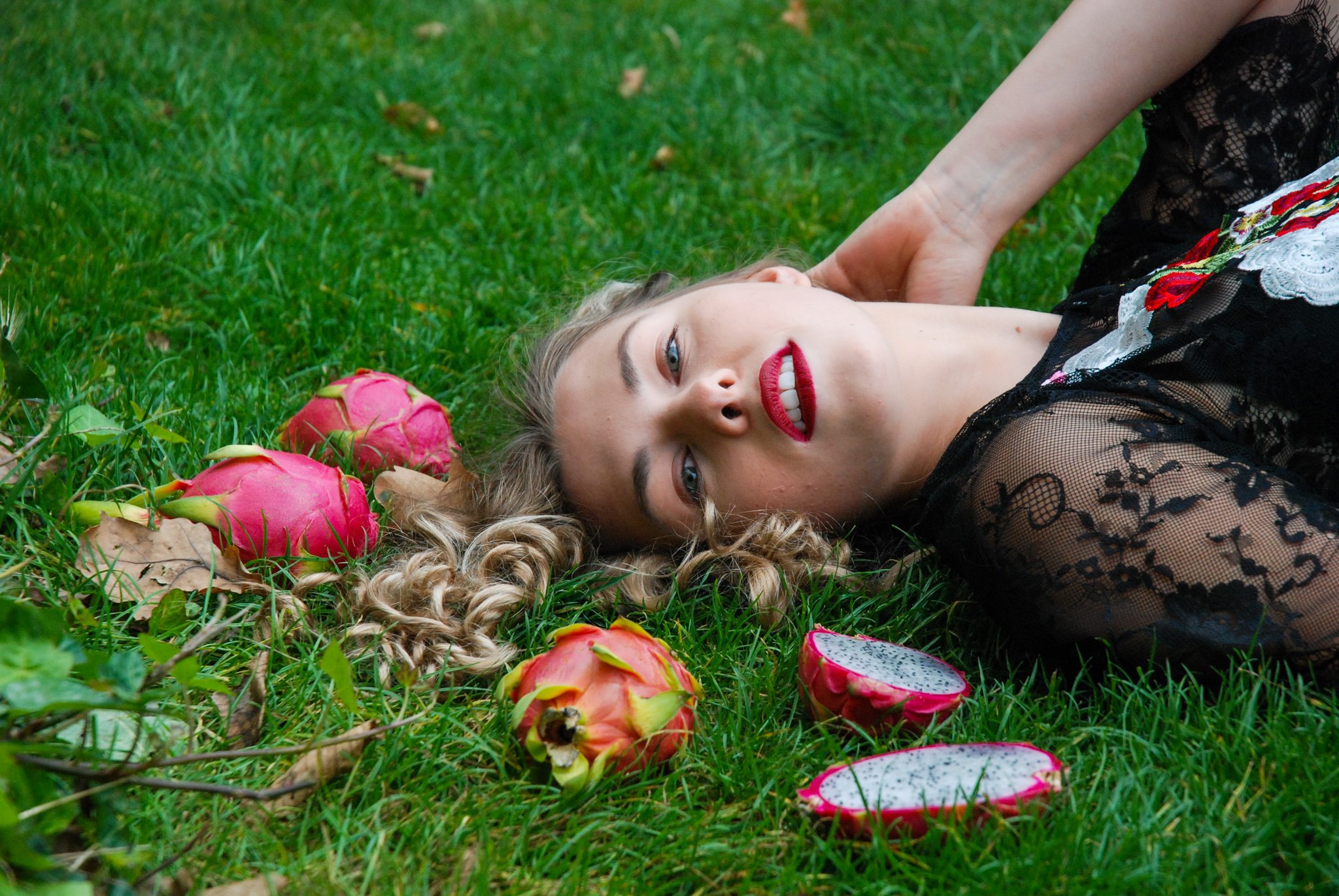 A woman with curly blonde hair lying on green grass surrounded by dragon fruit and leaves, wearing a black lace dress and red lipstick.