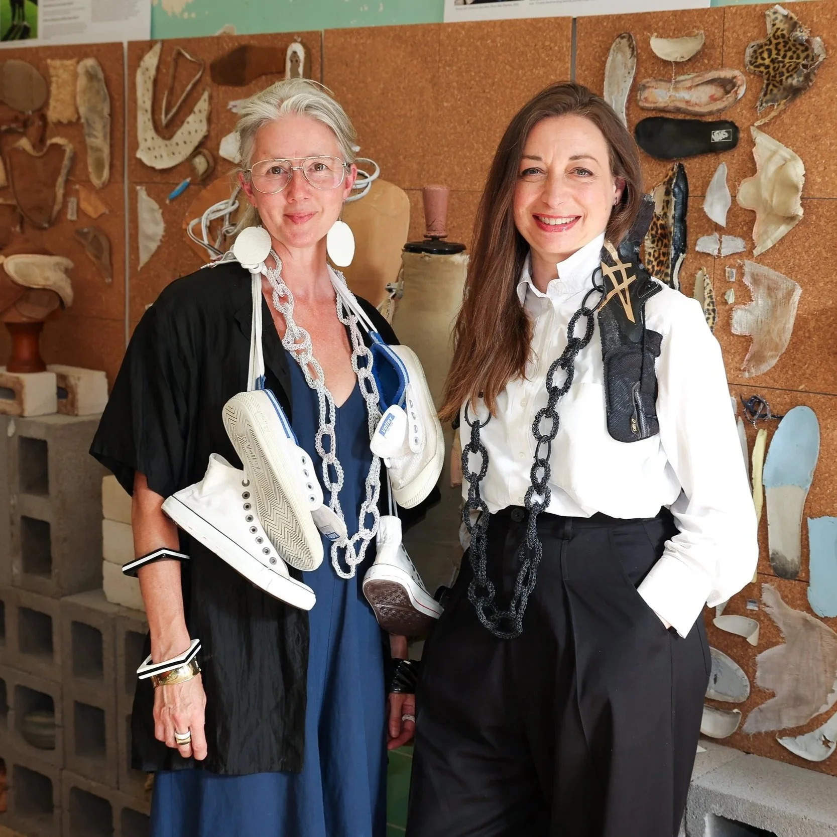 Pennie Jagello and Alexandra Sherlock standing in front of a wall of deconstructed shoes pinned in flat-lay formation. Pennie wears a pair of sneakers around her neck and Alex wears a black waistcoat made from a pair of black Asics sneakers