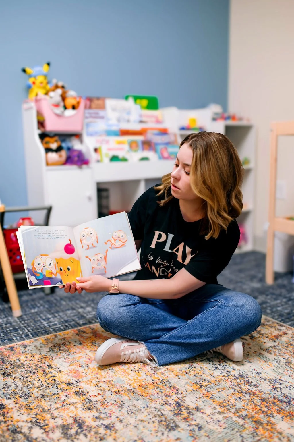 sitting cross-legged on a rug in a children's playroom, reading a colorful picture book about donuts and play therapy.