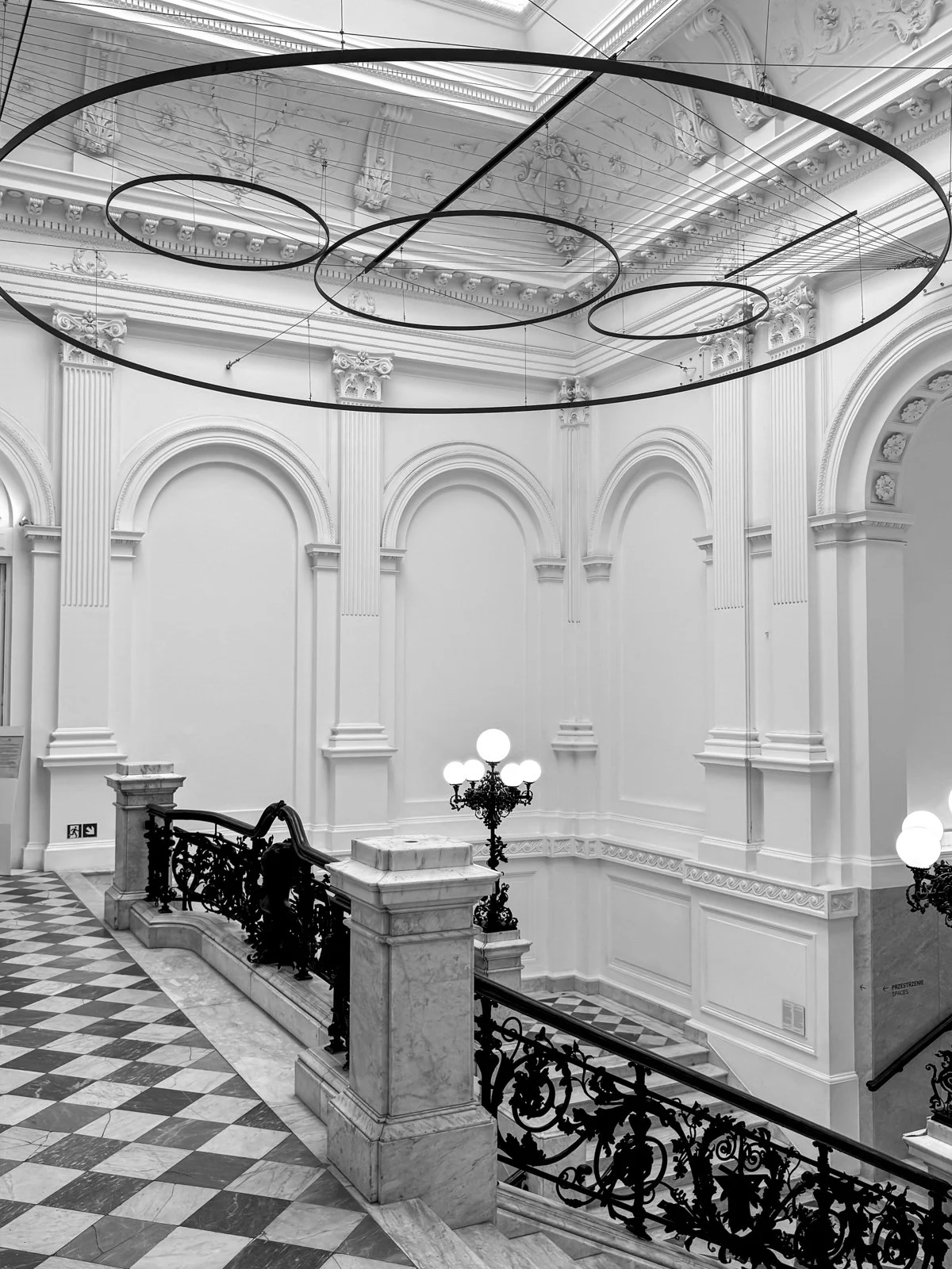Elegante, hohes Innere eines klassischen Gebäudes mit kunstvollen Wand- und Deataillierungen, großen runden Leuchten an der Decke, einem kunstvollen schwarzen Geländer, einer Marmortreppe und einer stilvollen Stehleuchte.