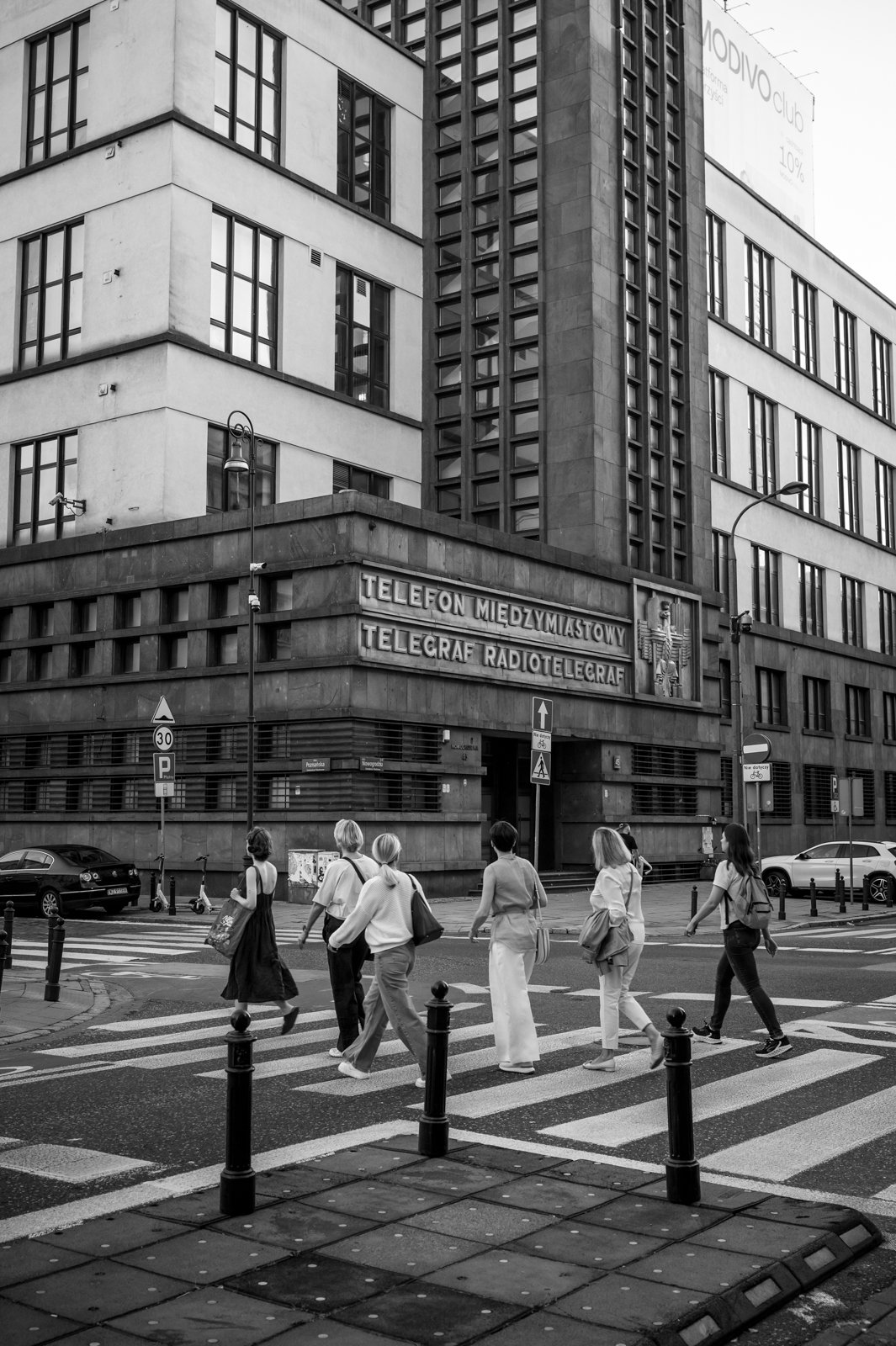 Schwarz-Weiß-Foto einer Stadtstraße mit mehreren Fußgängern, die den Zebrastreifen überqueren, vor einem großen modernen Gebäude mit Schildern in polnischer Sprache, die Telefondienst, Telegrafie und Radiotelegraphie bezeichnen.