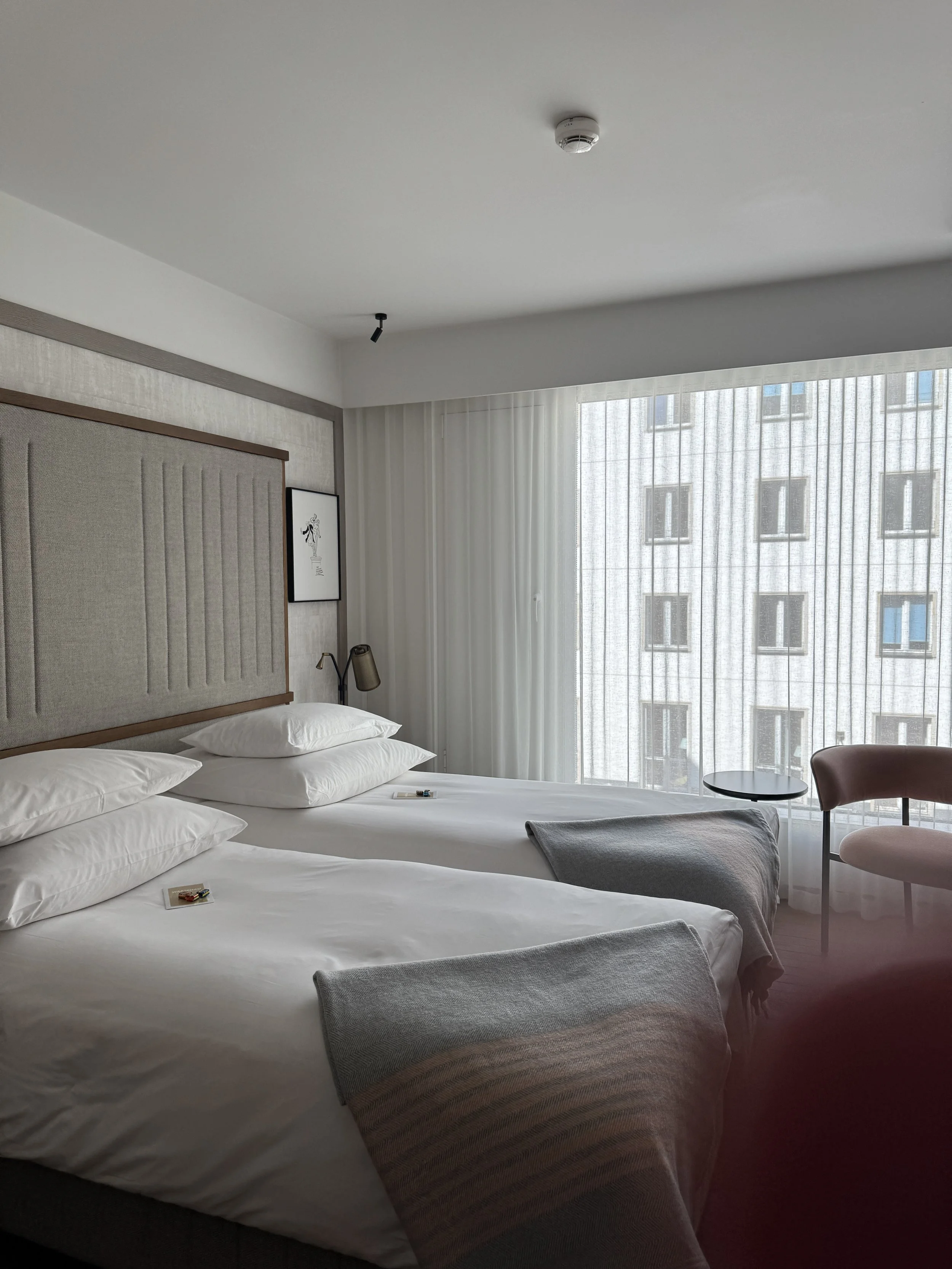 Ein modernes Hotelzimmer mit zwei Betten, einer Nachttischlampe, einem Sessel, einem kleinen Tisch und großen Fenstern mit transparenten Vorhängen, durch die ein Gebäude außerhalb sichtbar ist.