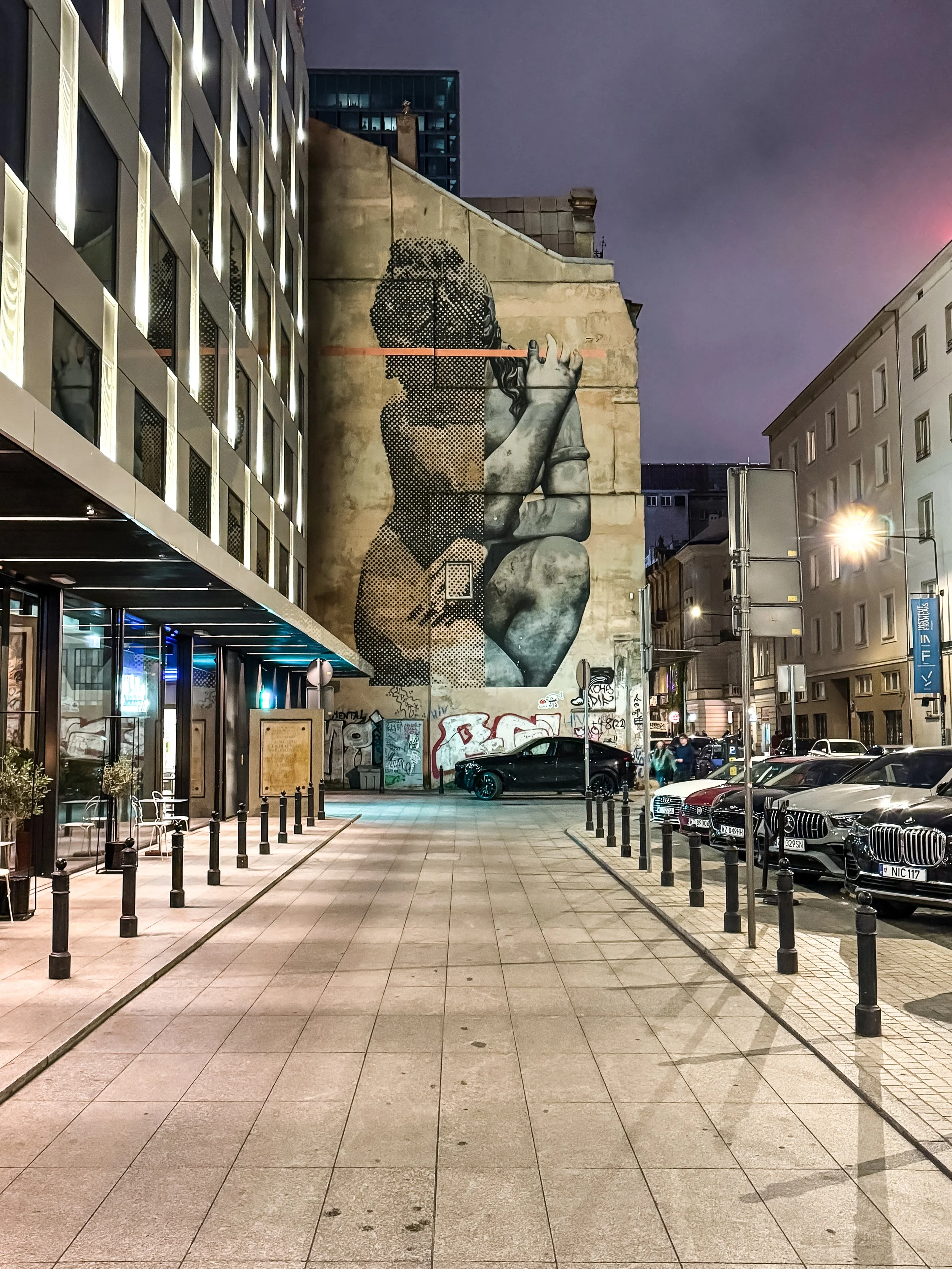 Straße in der Stadt bei Nacht mit einem großen Wandbild eines Mädchens, das in eine Richtung schaut, und einer Reihe geparkter Autos auf der rechten Seite.