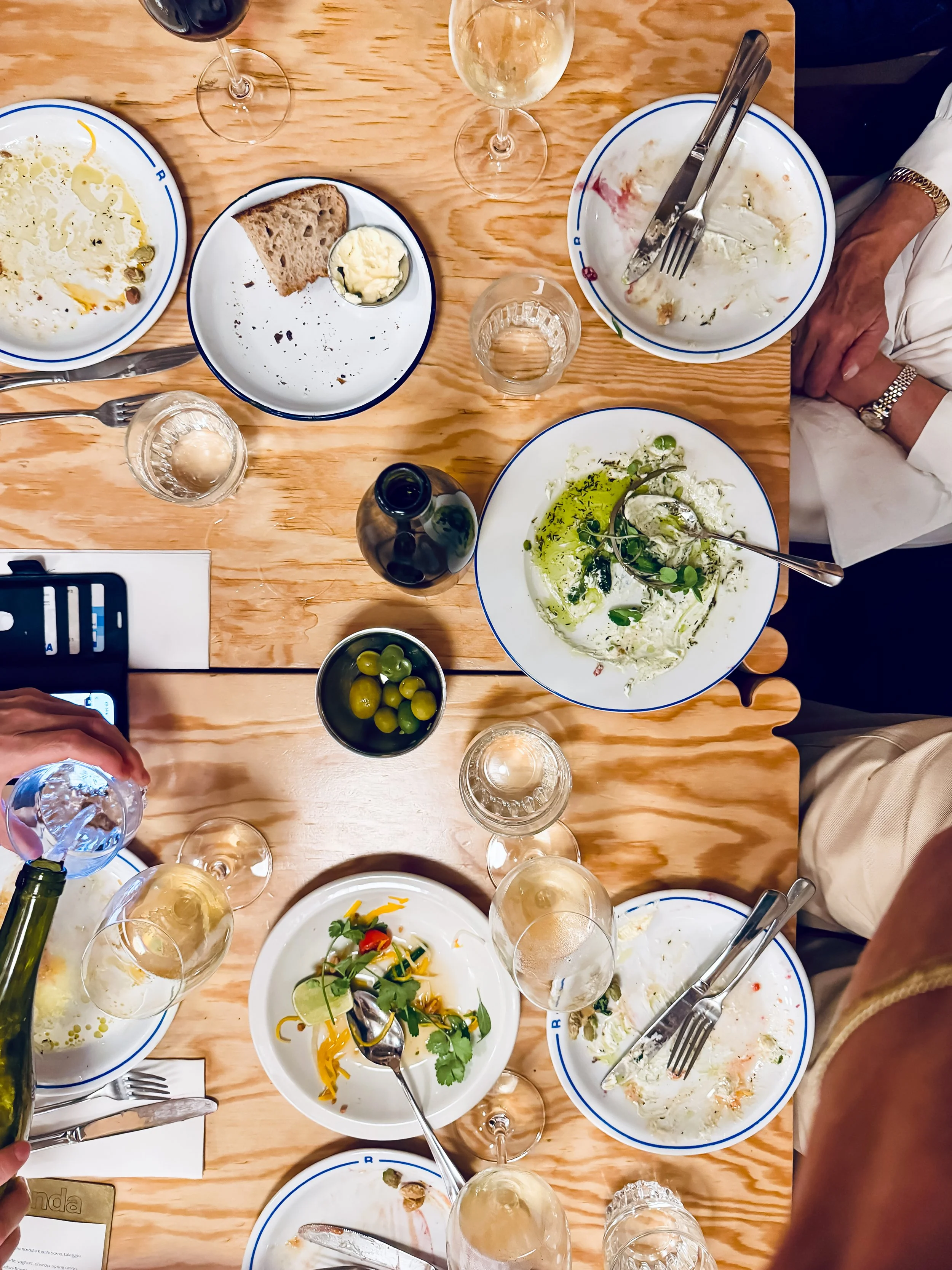 Abendessen auf einem Holztisch mit leeren Tellern, Gläsern und Besteck, zudem Essensreste und eine Flasche.