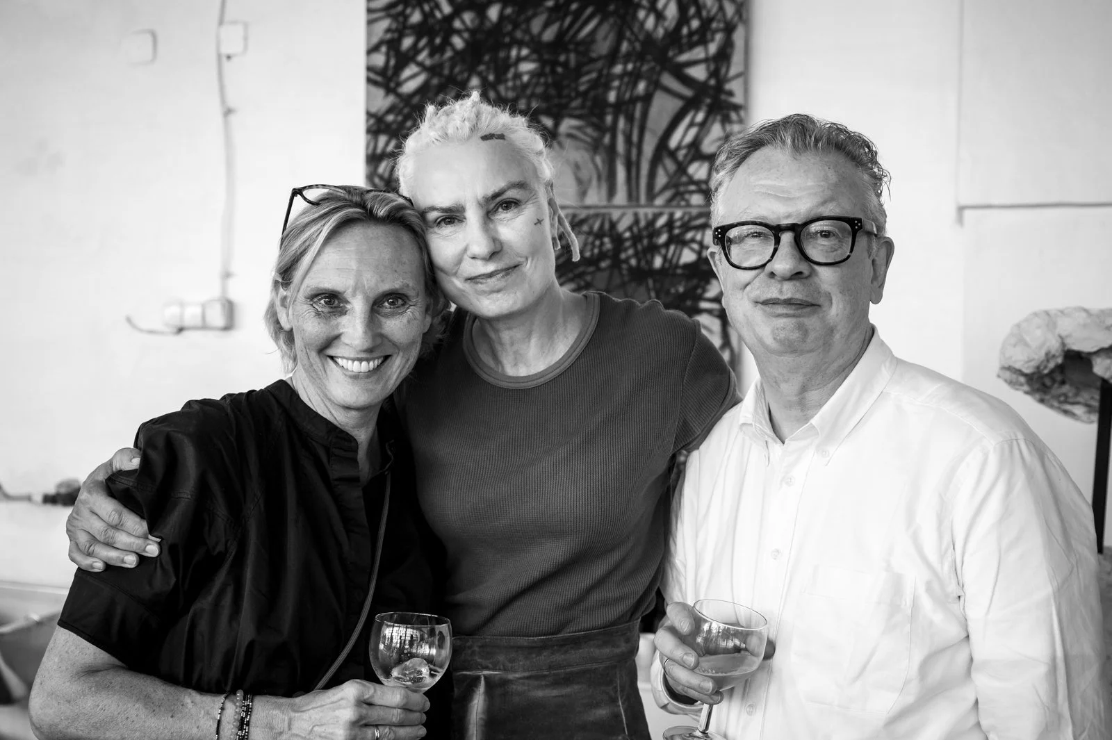 Drei Menschen, zwei Frauen und ein Mann, bei einer gesellschaftlichen Veranstaltung, lachen und halten Gläser Wein in der Hand. Hintergrund mit Kunstwerken.