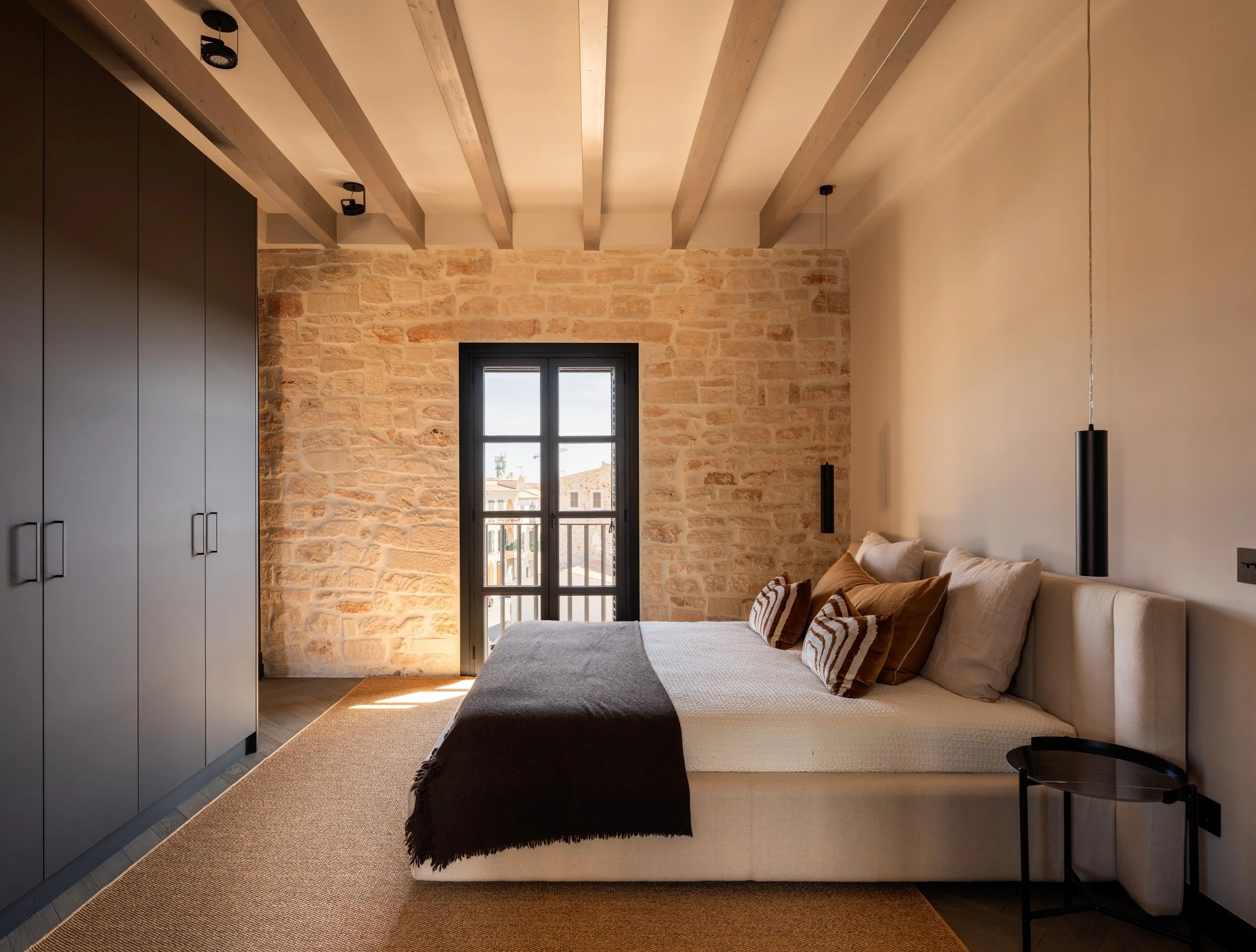 Schlafzimmer mit einem großen Bett, schwarzen Vorhängen vor einem Fenster, einer gestrickten Decke, beige Kissen, einem roten Kissen, einem schwarzen Nachttisch, einer weißen Wand, einem schwarzen Lampenschirm, einer Wandlampe, Holzpaneelen an der Decke, einer Steinwand und einem beige Teppich.