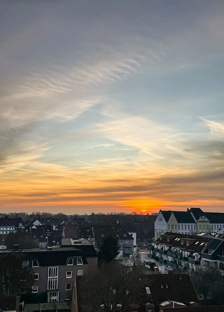 Sonnenuntergang über einer Stadt mit mehrstöckigen Häusern, Wolken und einem farbigen Himmel.