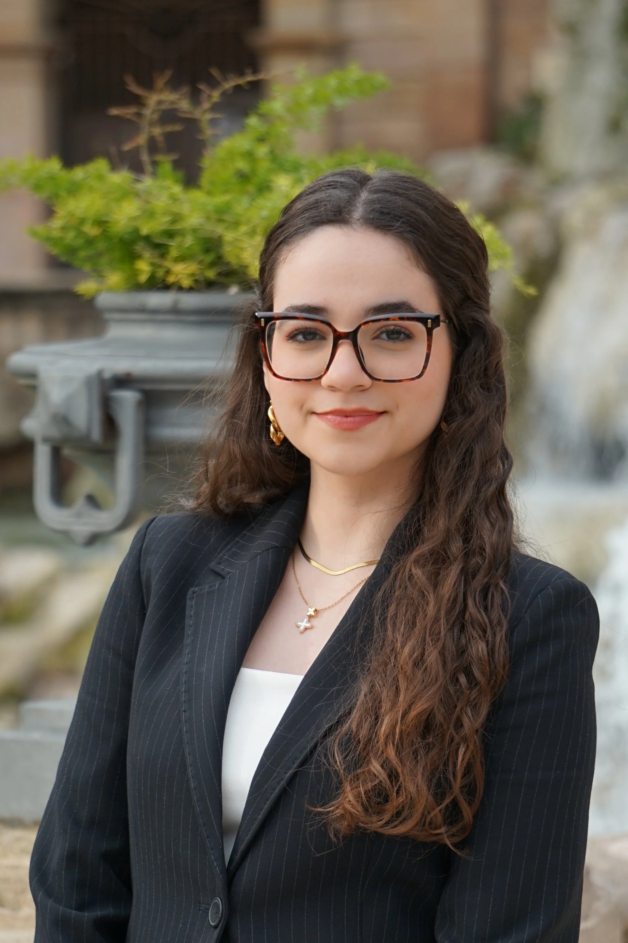 Joven mujer con gafas, cabello rizado y largo, usando saco negro y blusa blanca, posando frente a una fuente con plantas verdes y un fondo de piedra y estructura.