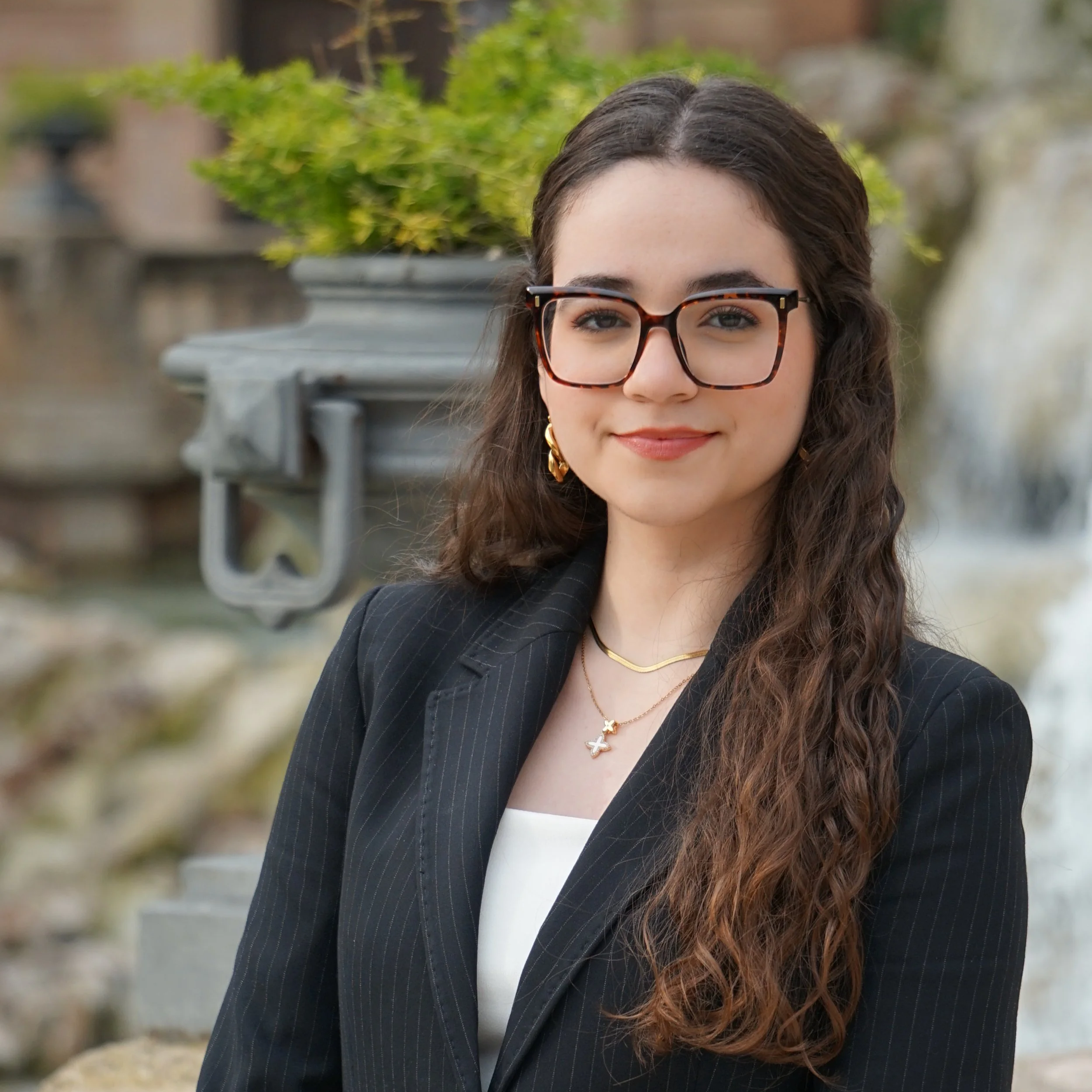 Retrato de una joven mujer con cabello castaño y piel clara, vestida con una blusa blanca de botones y un blazer negro, sonriendo, en un entorno exterior con fuente y edificio antiguo en el fondo.