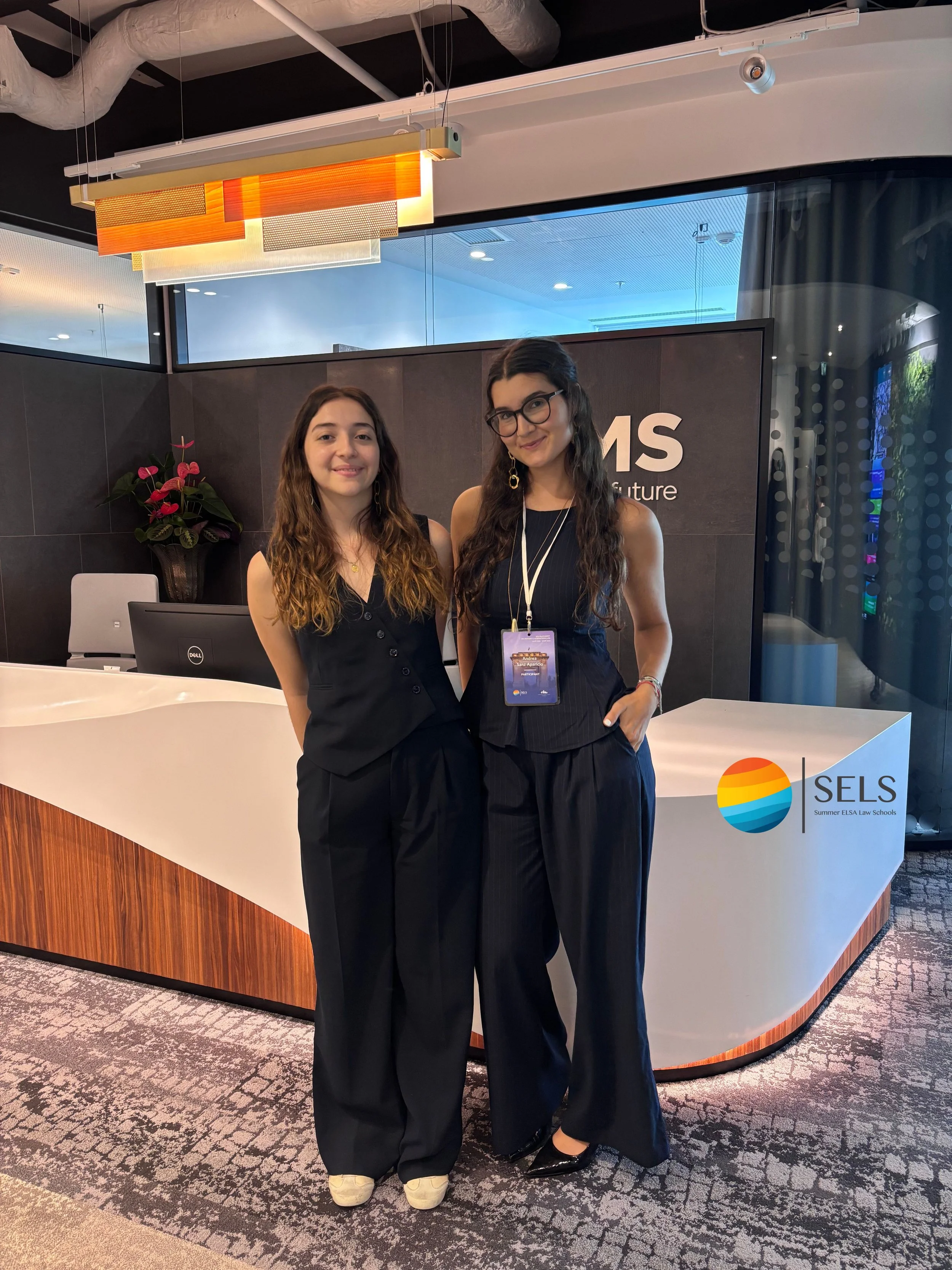 Dos mujeres con vestimenta formal en un mostrador de recepción de un edificio o evento, con logotipo de SELS en el fondo y decoraciones en el interior.