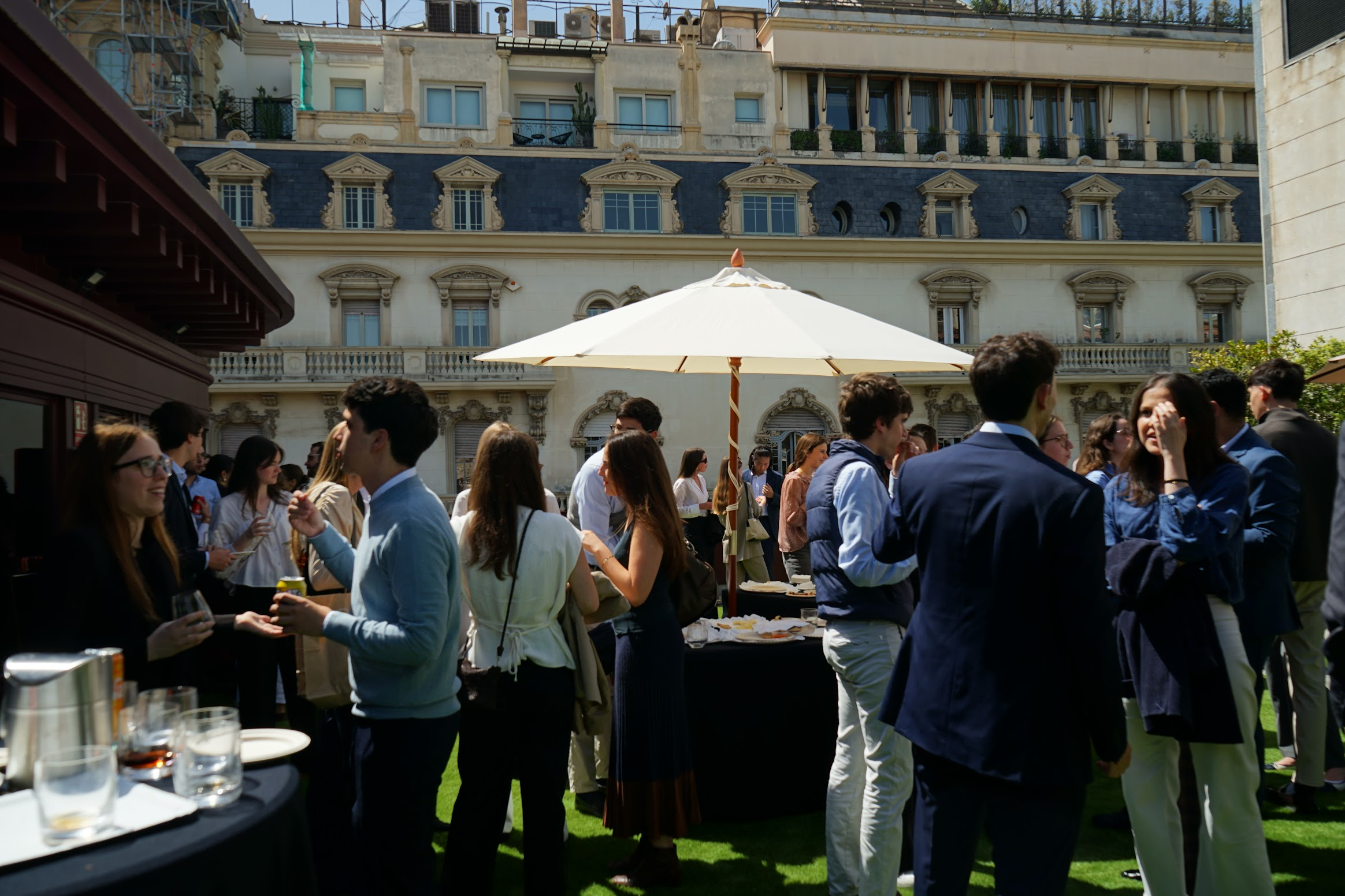 Grupo de personas en una reunión al aire libre con un edificio antiguo de fondo, hay una sombrilla grande y una mesa con bebidas y comida.