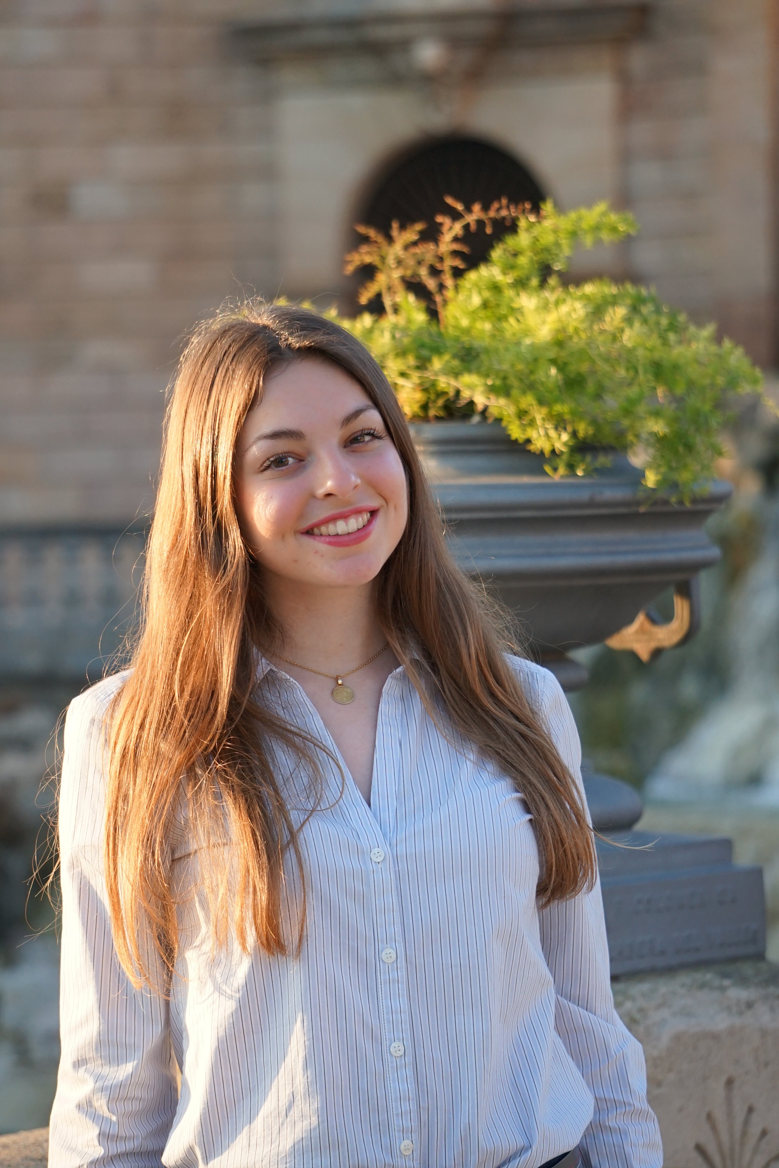 Joven mujer con cabello largo y rubio, sonriendo y vistiendo una blusa de rayas blancas y azules, en un lugar al aire libre con una fuente y plantas verdes al fondo.