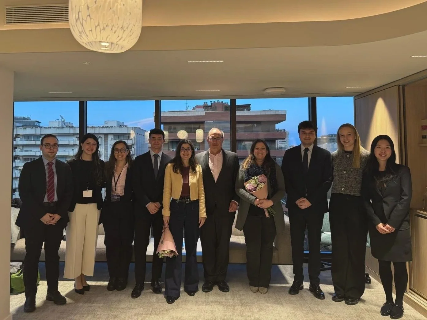 Grupo de personas en una reunión, con fondo de ventanas que muestran edificios urbanos.