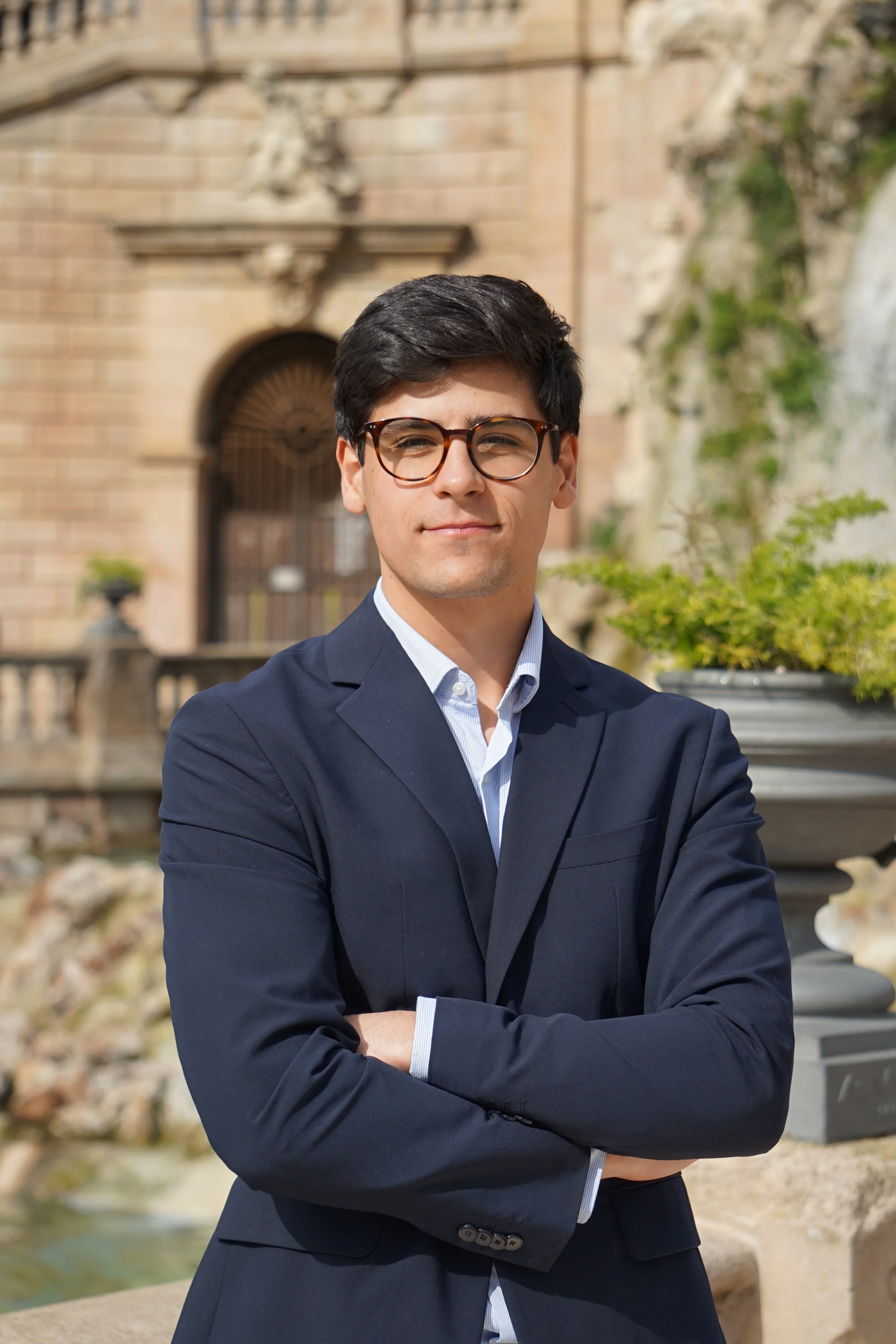 Hombre con gafas, traje oscuro y camisa blanca, de pie con brazos cruzados frente a un edificio de piedra y un jardín en un día soleado.