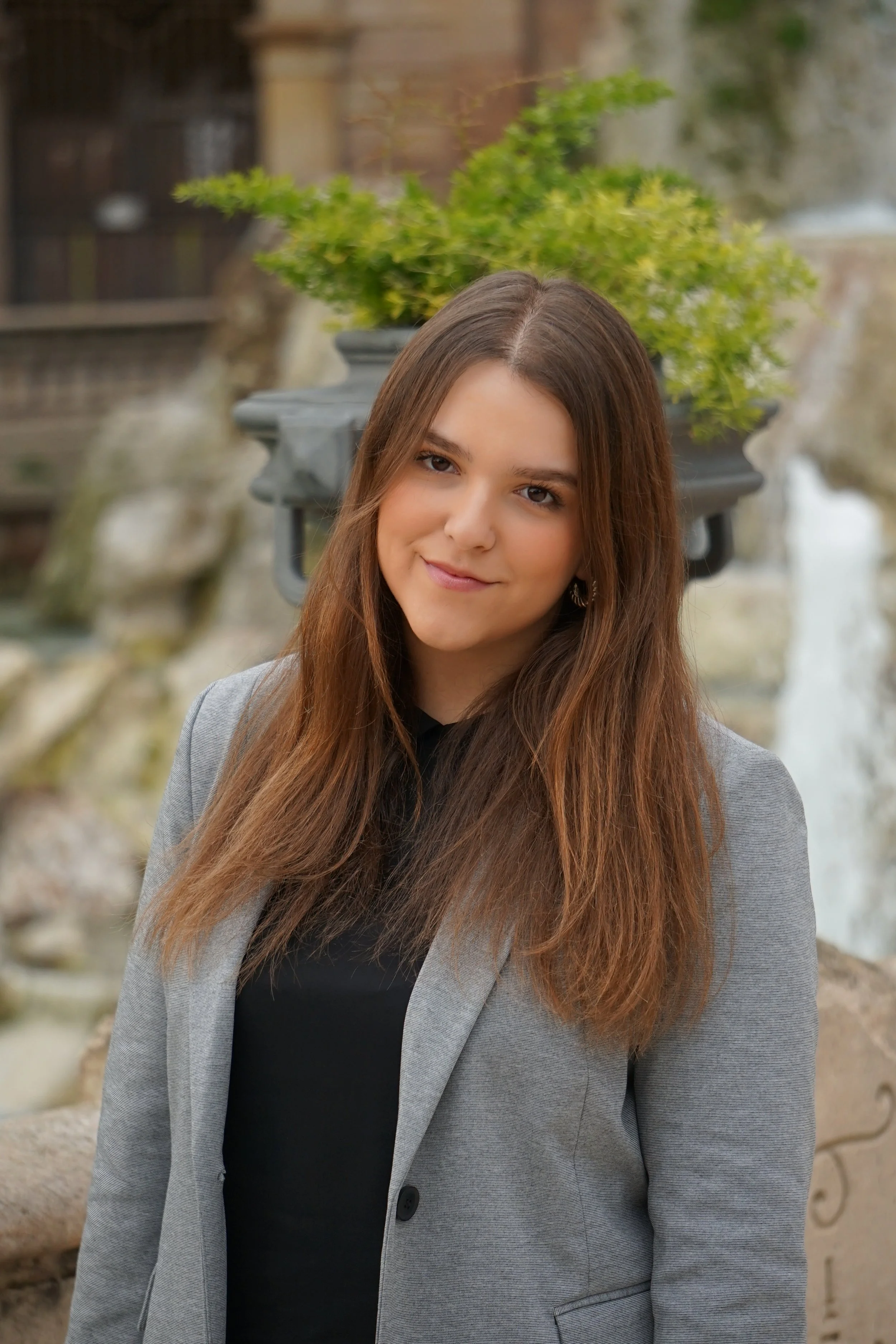 Mujer joven con cabello largo y liso, lleva un saco gris y una blusa negra, sonríe ligeramente y está en un entorno al aire libre con una fuente y plantas en el fondo.