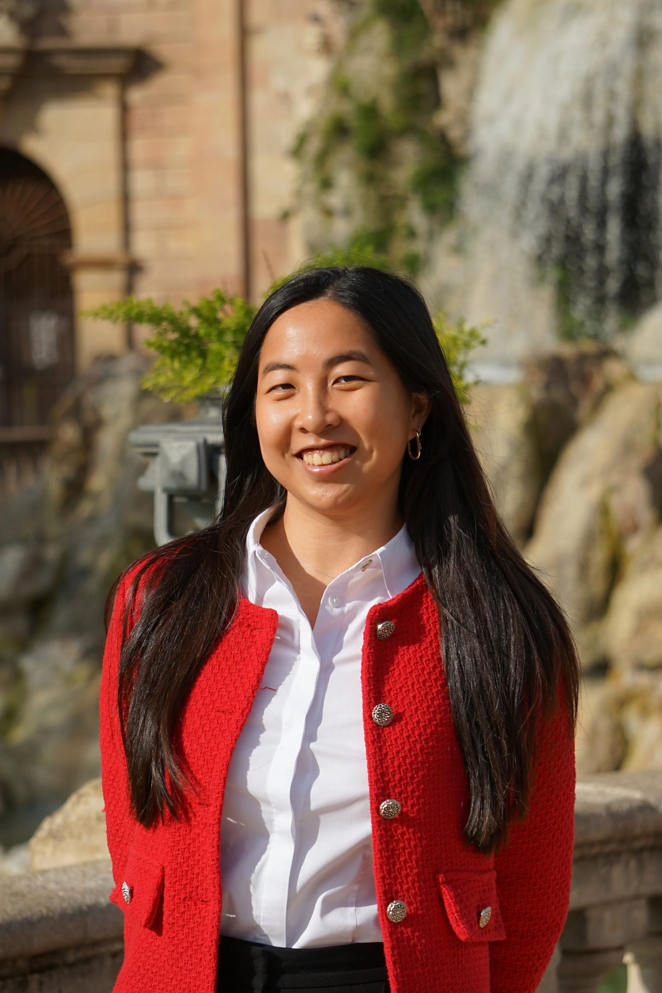 Mujer sonriendo con fondo de agua y naturaleza, vestida con blusa blanca y suéter rojo.