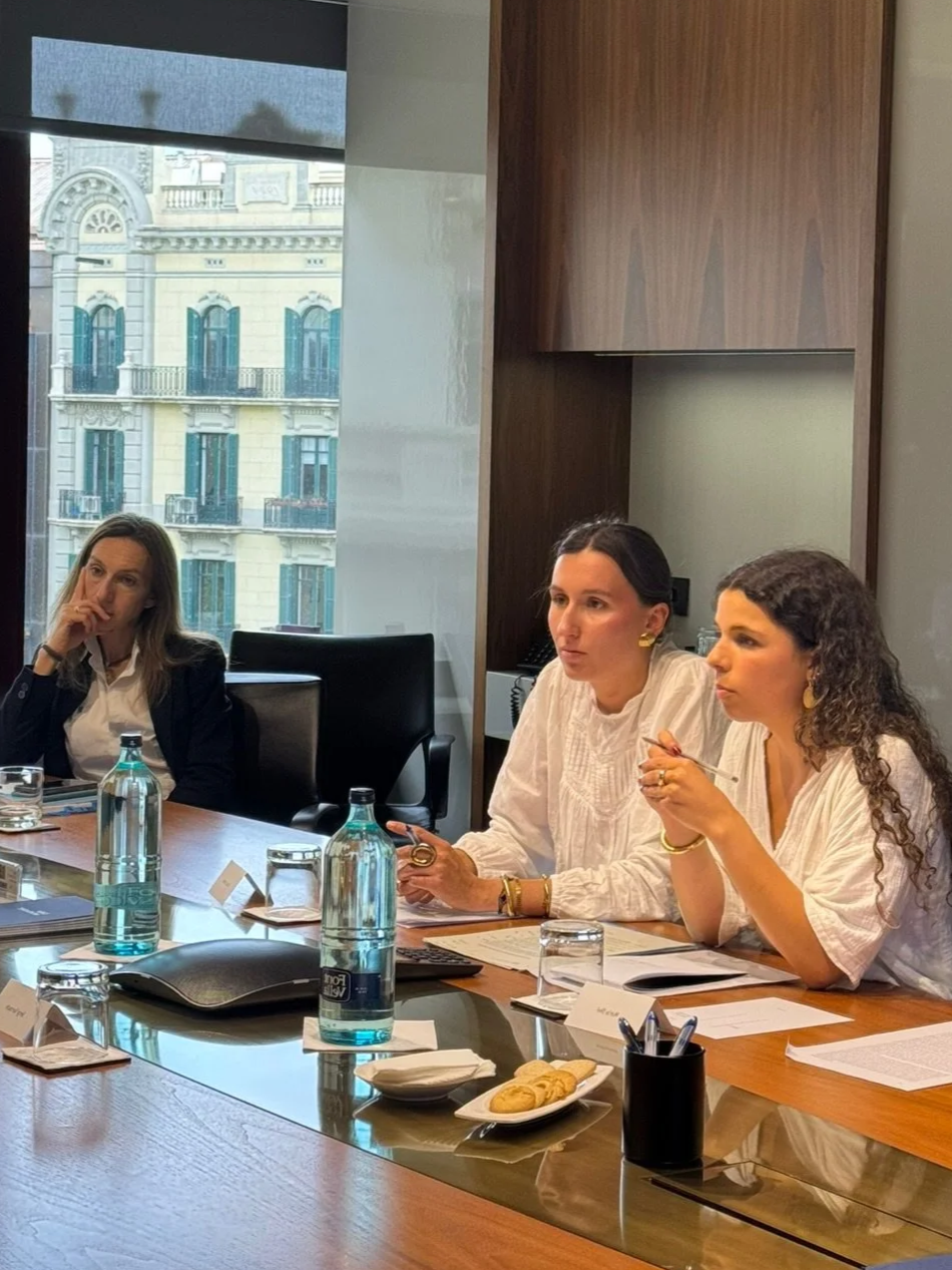 Tres mujeres sentadas en una mesa de conferencia discutiendo, con documentos, botellas de agua, y una sala de oficinas con ventanas y edificios en el fondo.