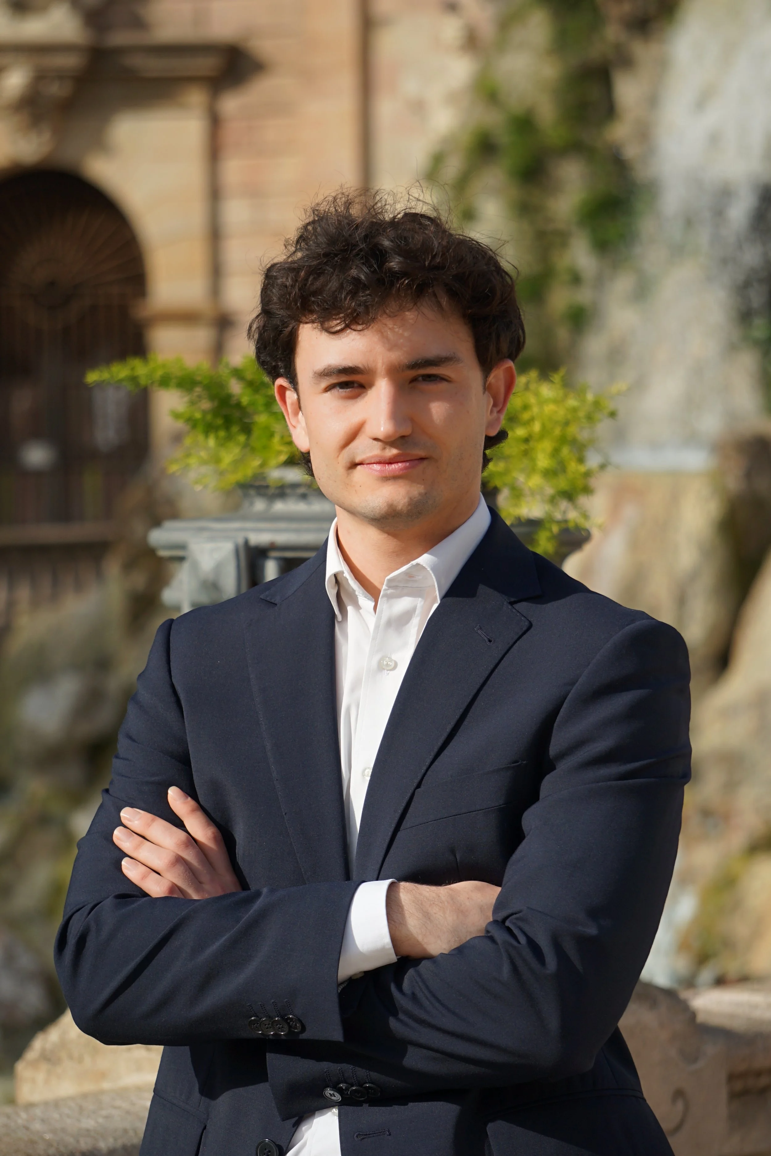 Joven en traje oscuro posando con brazos cruzados en un lugar exterior con fuente y vegetación.