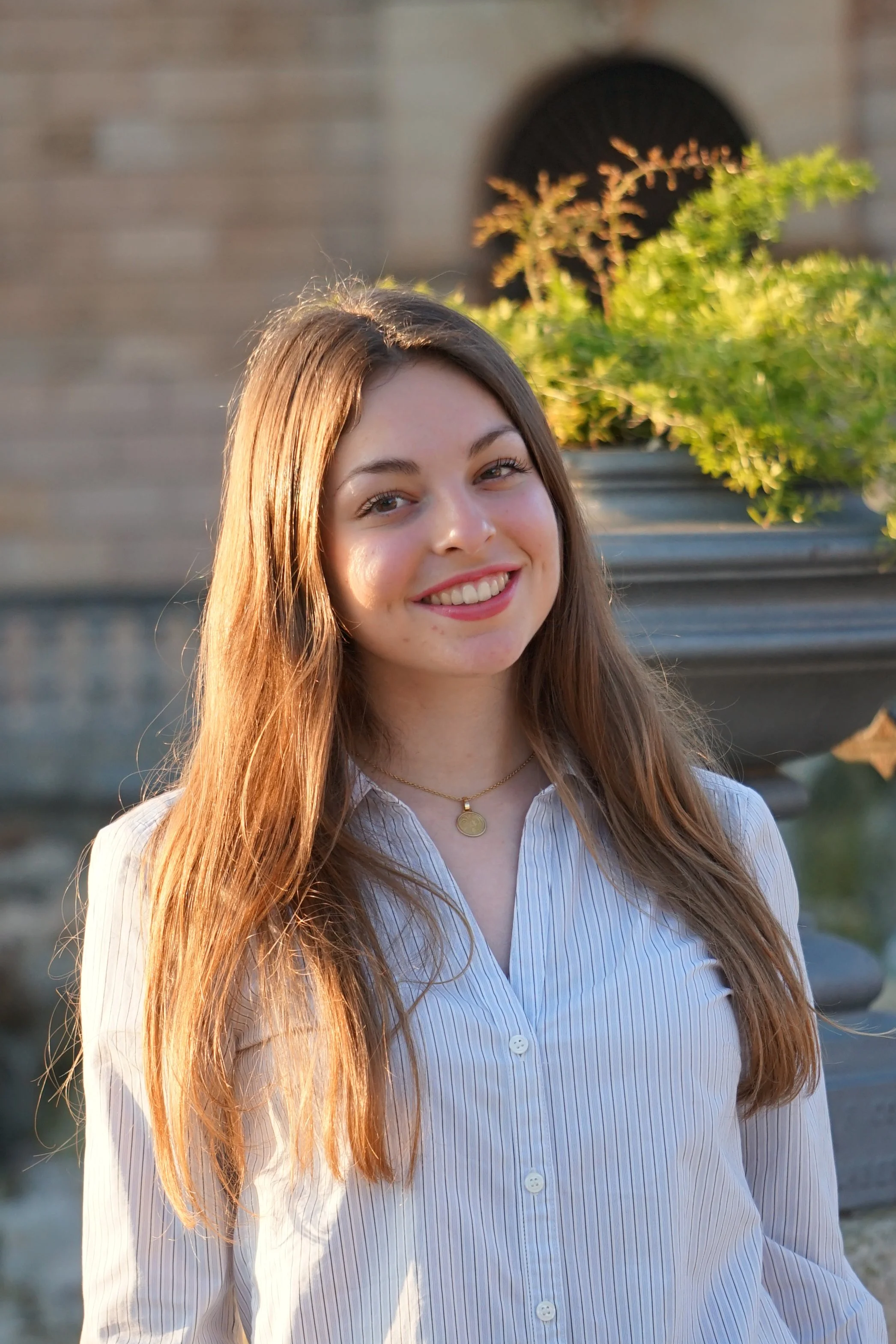 Joven mujer con cabello largo y rizado, sonrisa, camisa blanca a rayas, en ambiente exterior con plantas y estructura de ladrillo en el fondo, luz natural del atardecer.