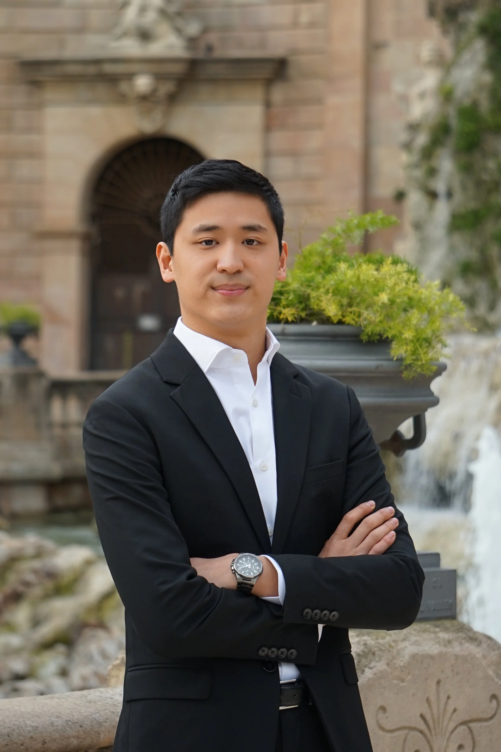 Hombre joven en traje negro y camisa blanca con brazos cruzados, de fondo una fuente y un edificio antiguo.