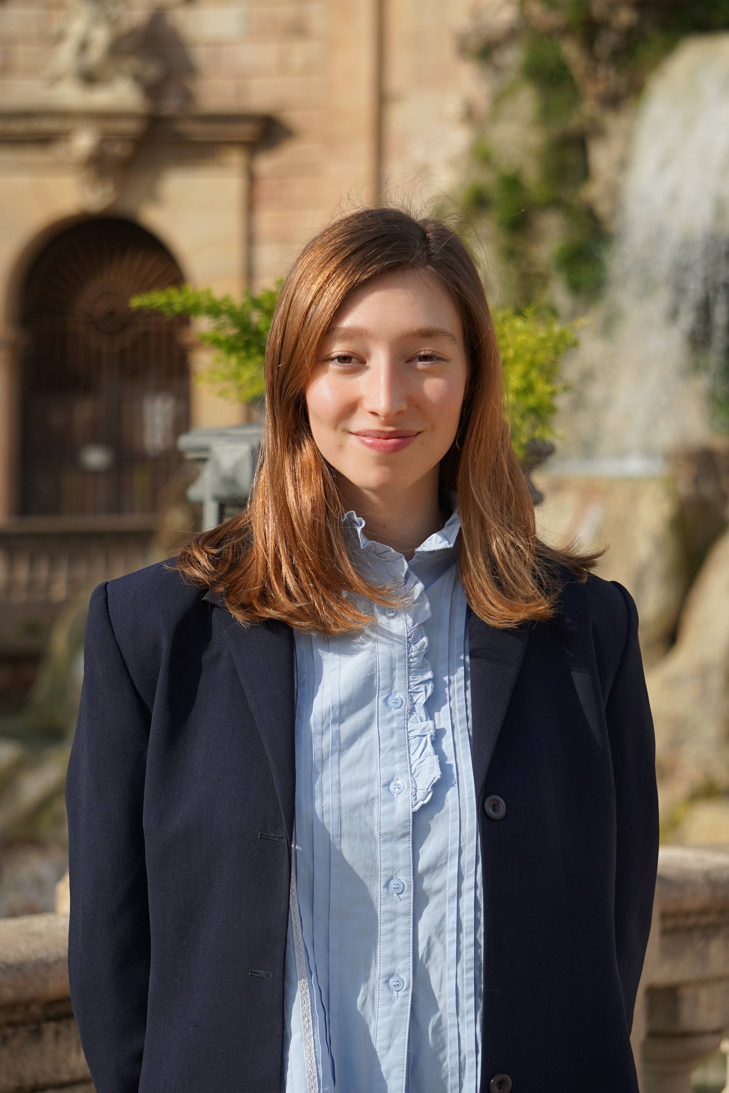 Retrato de una joven mujer con cabello castaño y piel clara, vestida con una blusa blanca de botones y un blazer negro, sonriendo, en un entorno exterior con fuente y edificio antiguo en el fondo.