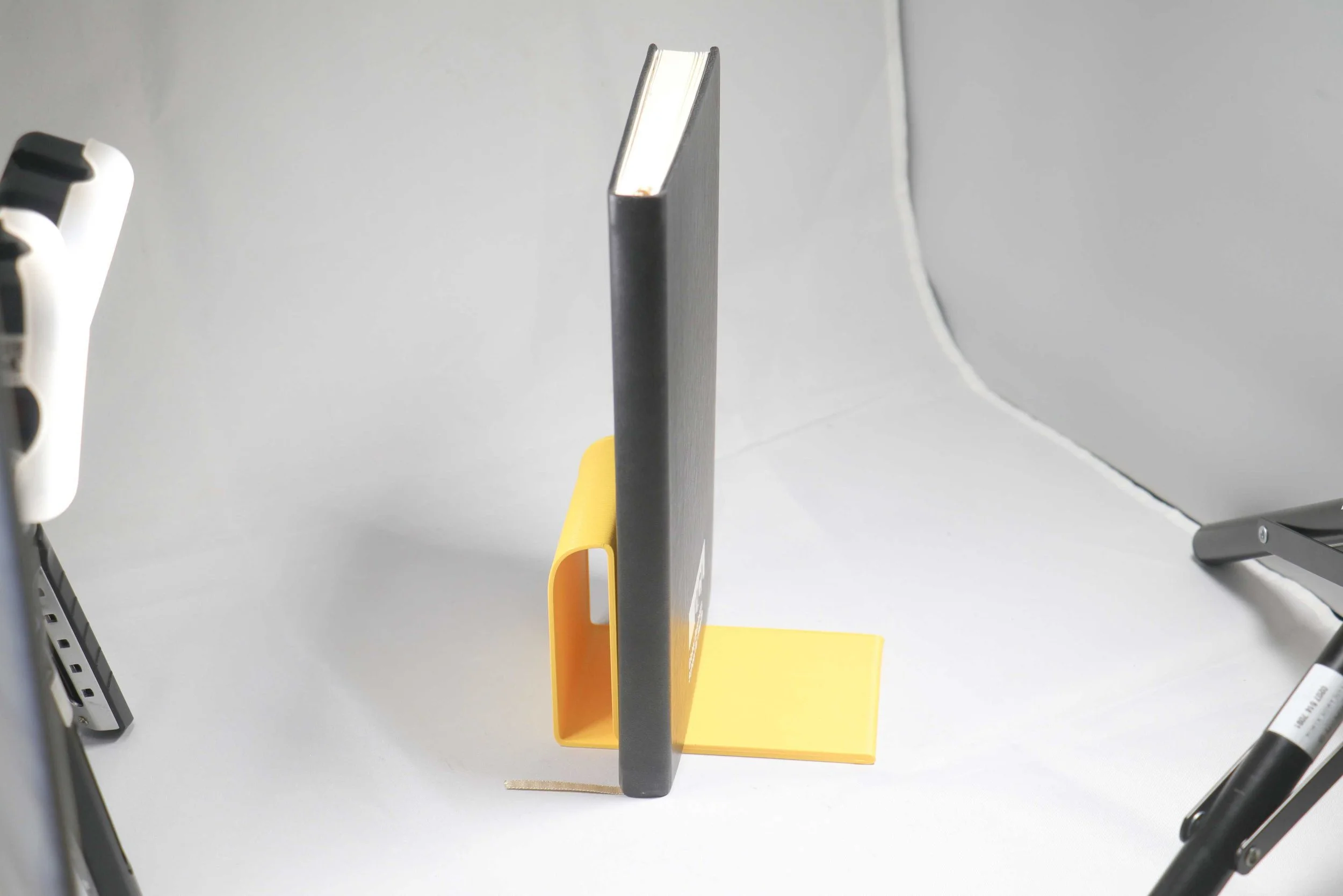 Black book or notebook placed upright in yellow book stand on white surface, with parts of various office supplies visible on the sides and background.