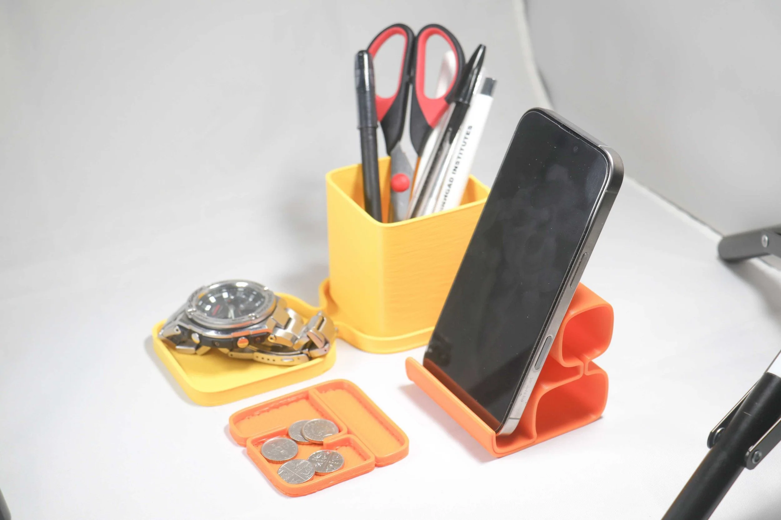 Desk with watch, coins in a case, and a smartphone in orange and yellow holders surrounded by pens and scissors in a yellow container.