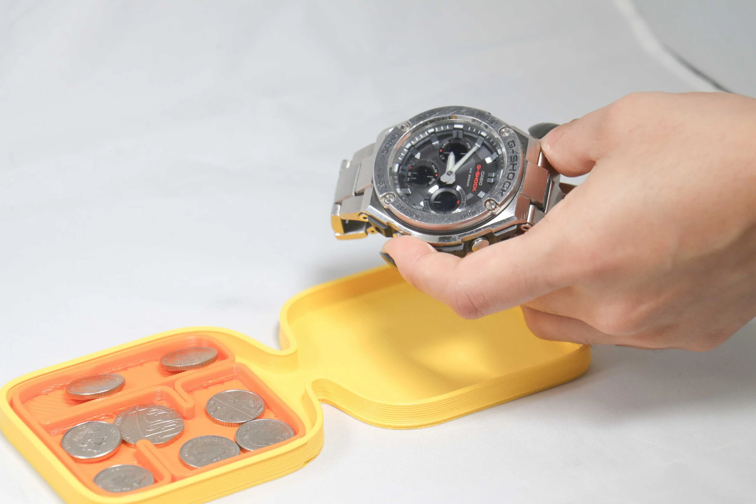 A person's hand holding a silver G-Shock watch over a yellow coin case with coins inside, on a white surface.