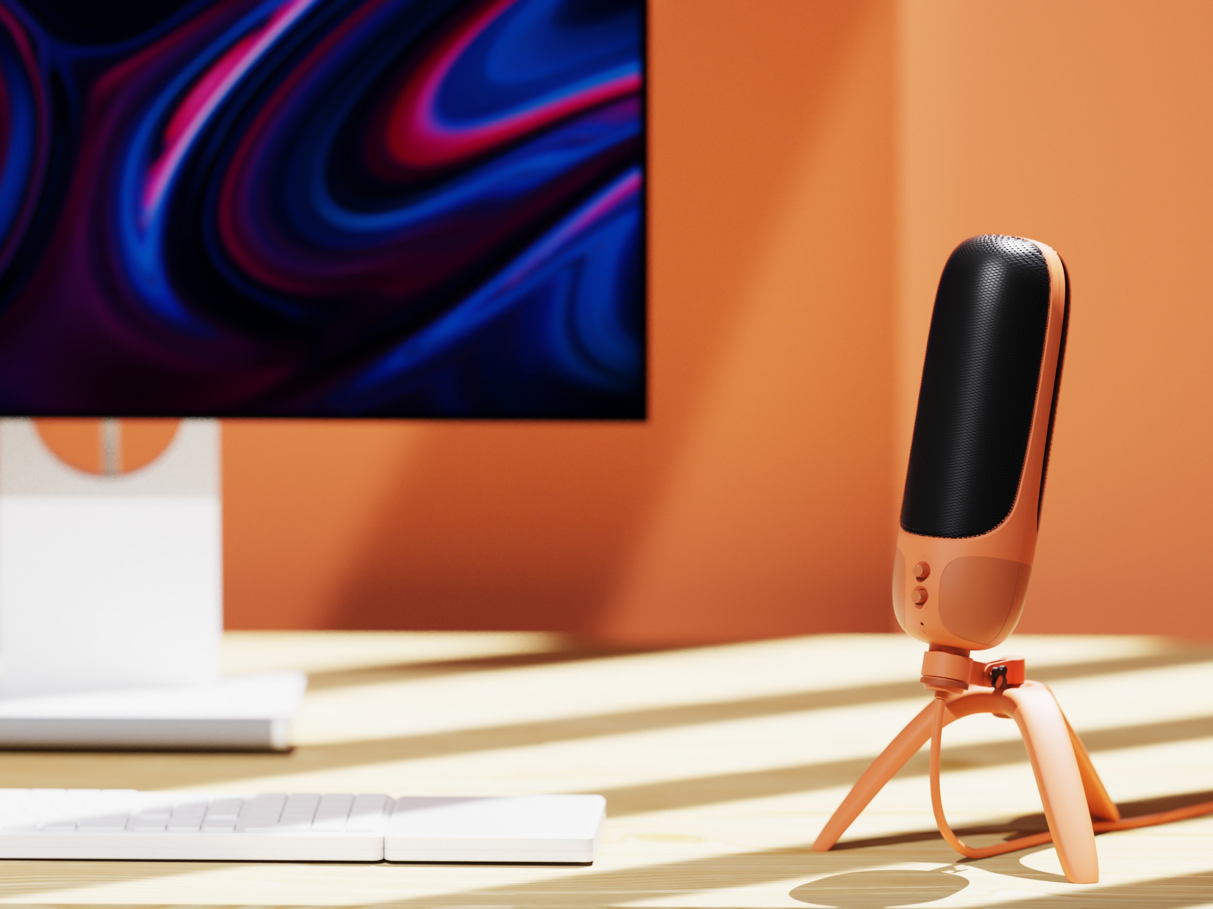 A microphone on a small tripod stand placed on a light-colored desk in front of a computer monitor with a colorful swirling pattern screen, against an orange wall background.