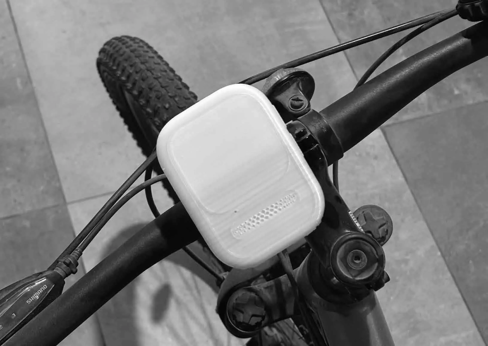 Black and white image of a bicycle's handlebar with a large, rectangular, white container attached to the center, a knobby tire, and visible cables.