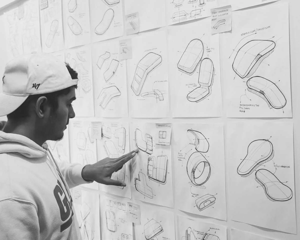 A person examining design sketches of product prototypes on a wall, including various sketches of ergonomic objects and device components.