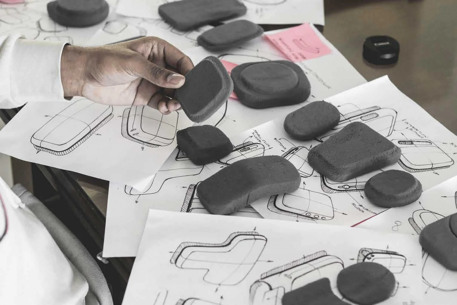 Person holding a foam piece above sketches and prototypes of a device on a table.
