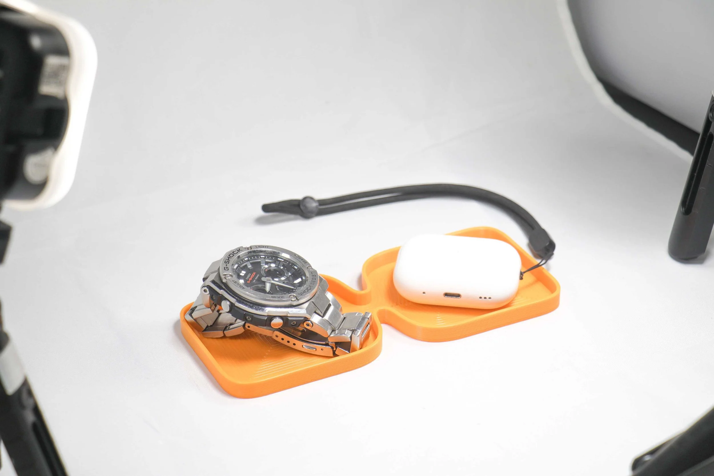 A silver wristwatch, a white wireless earbud case, and a black charging cable on orange surfaces against a white background.