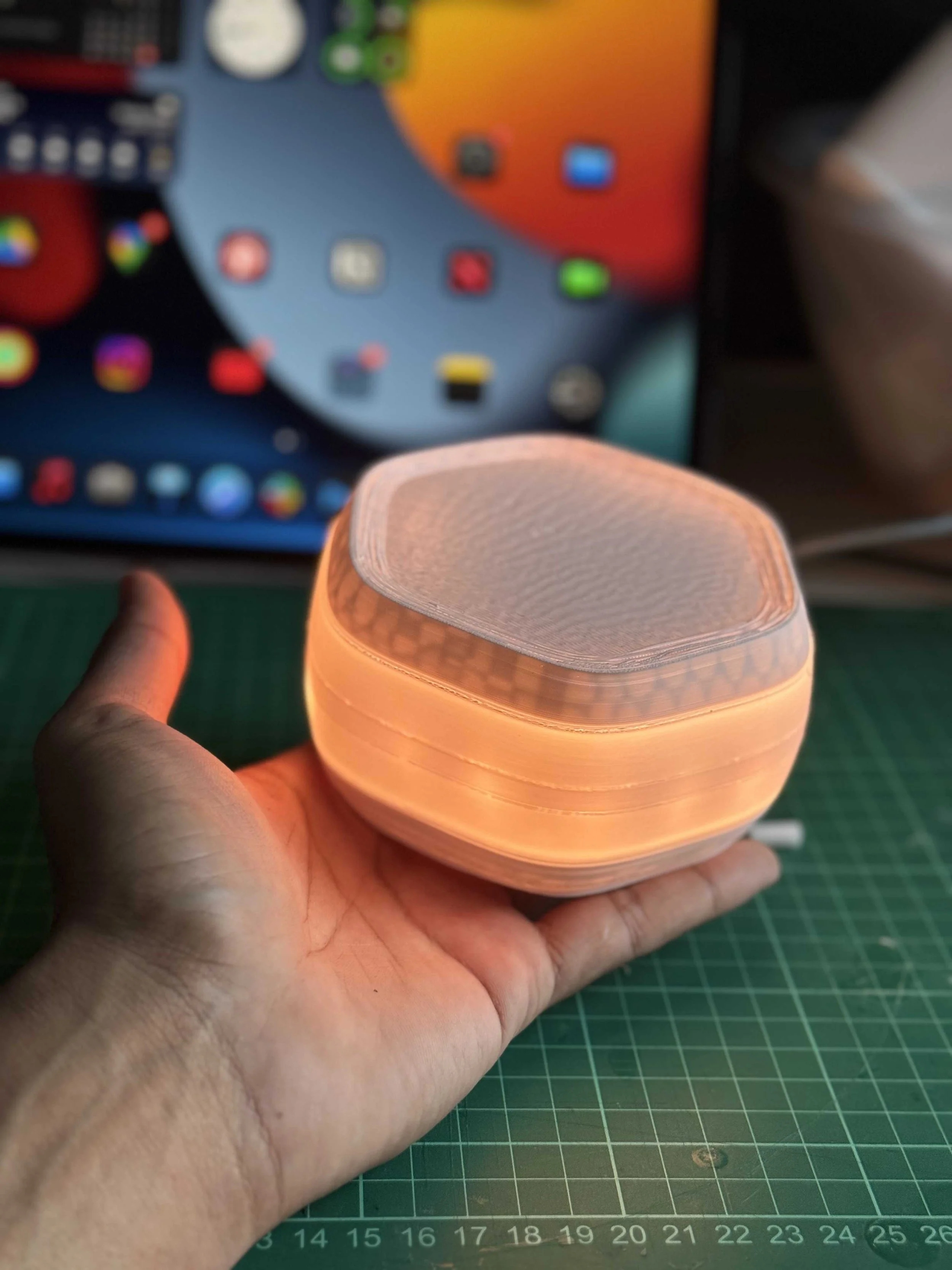 A person holding a glowing, round, wooden lamp with a hexagonal top in front of a tablet with a colorful screen.