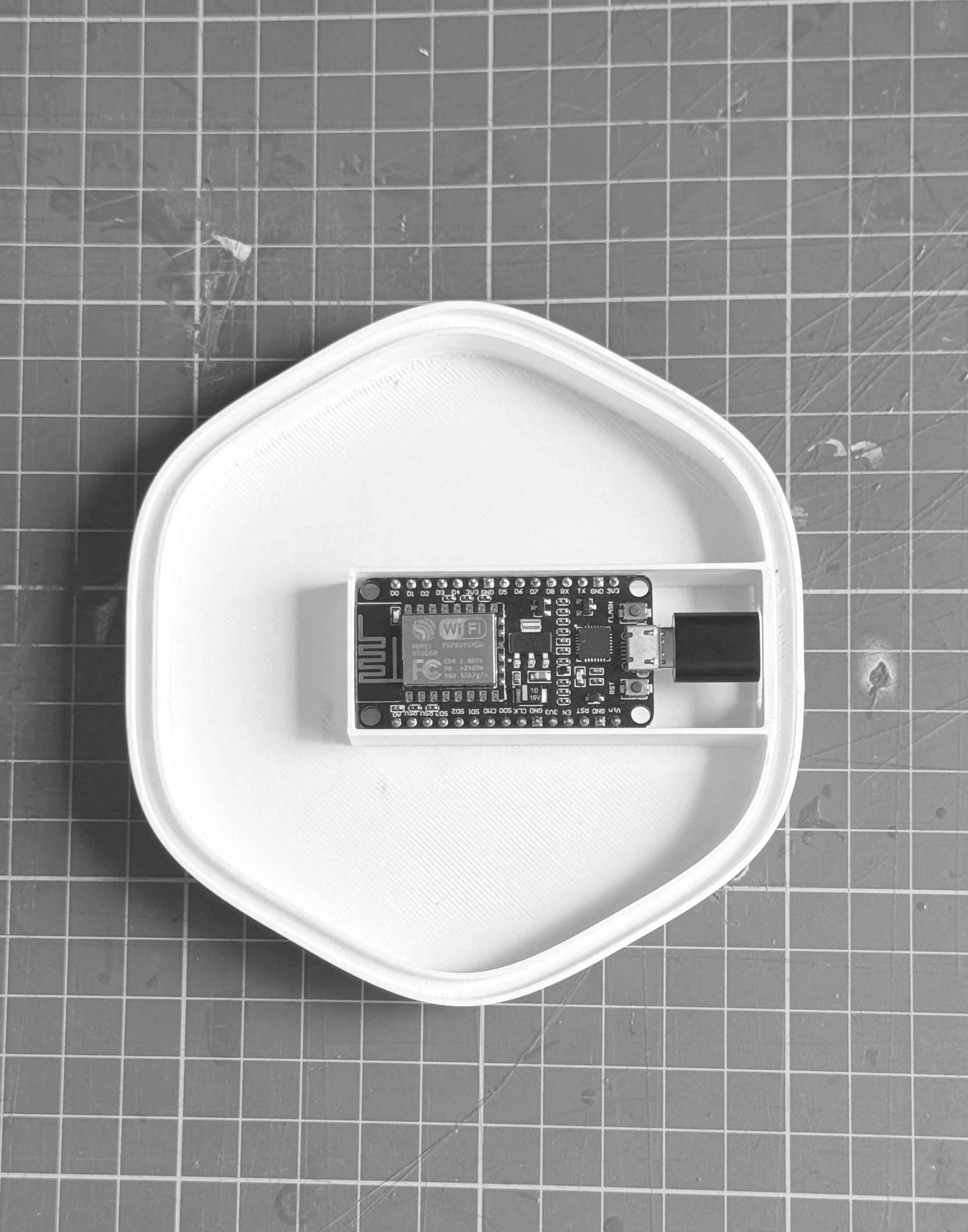 A small electronic microcontroller module placed inside a rectangular white container on a grid-patterned work surface.