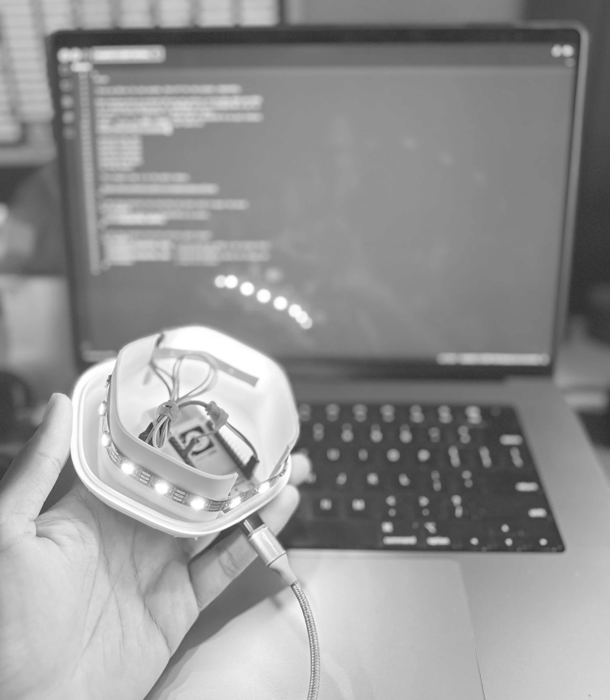 A person holding an LED circuit board with wiring and a USB cable in front of a computer monitor with code displayed.
