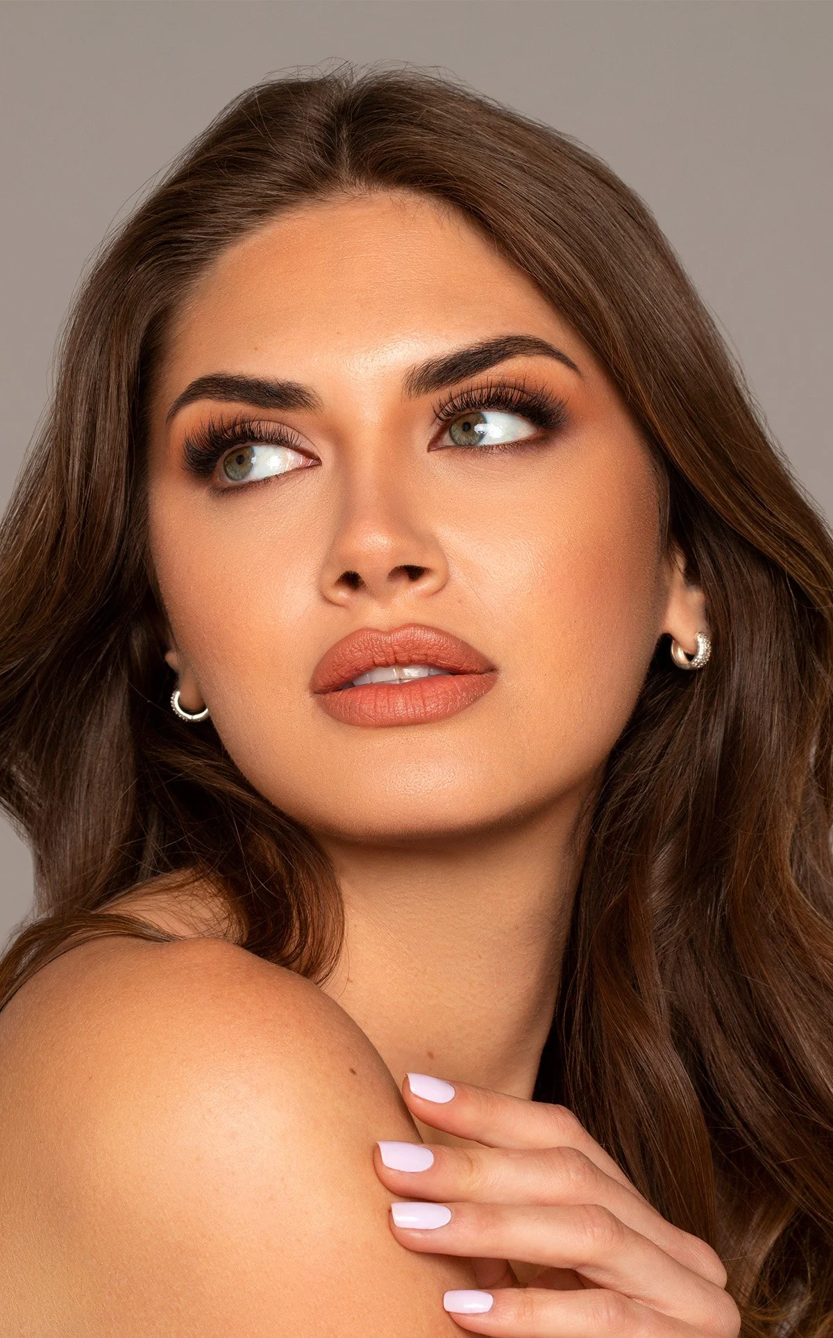 A close-up portrait of a woman with long, wavy brown hair, wearing makeup, earrings, and with her hand on her shoulder against a gray background.