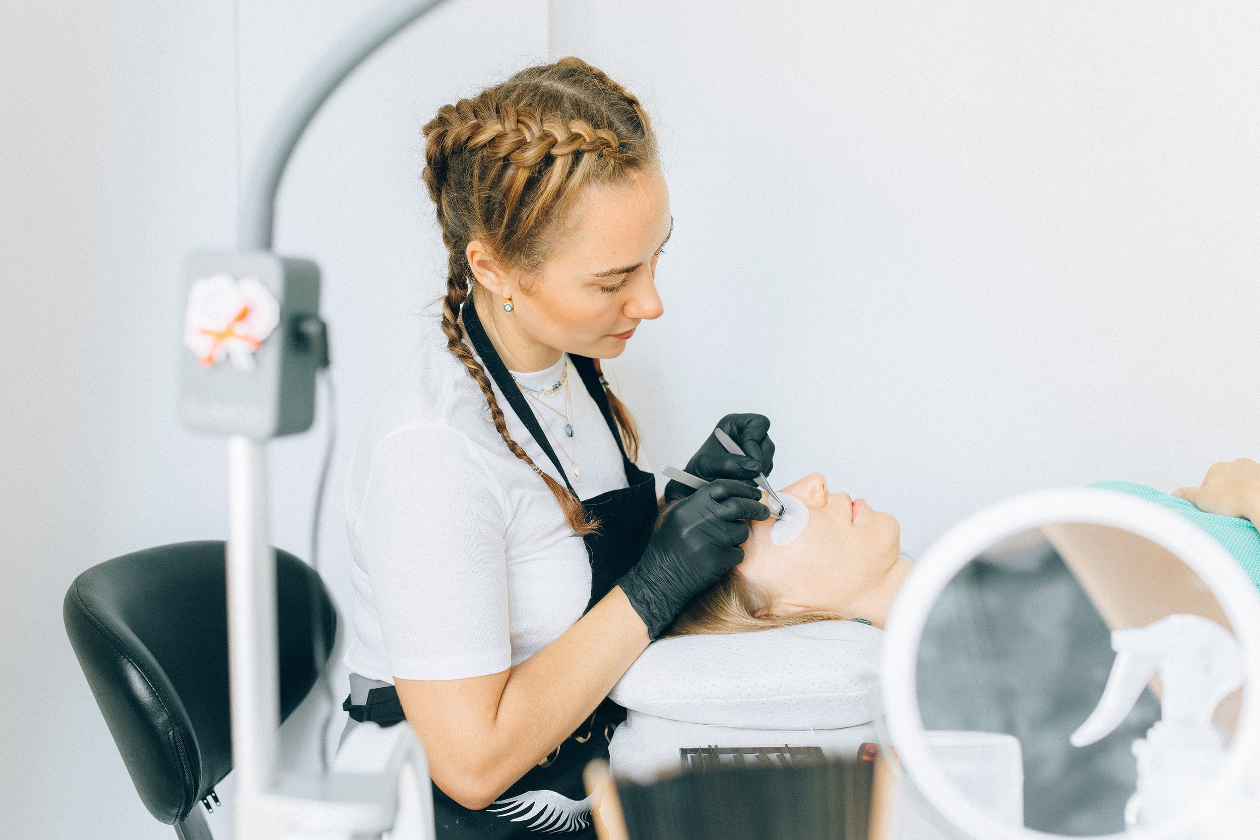 A woman with braided hair providing a cosmetic or skincare treatment to a woman lying down with closed eyes, in a clinical setting.