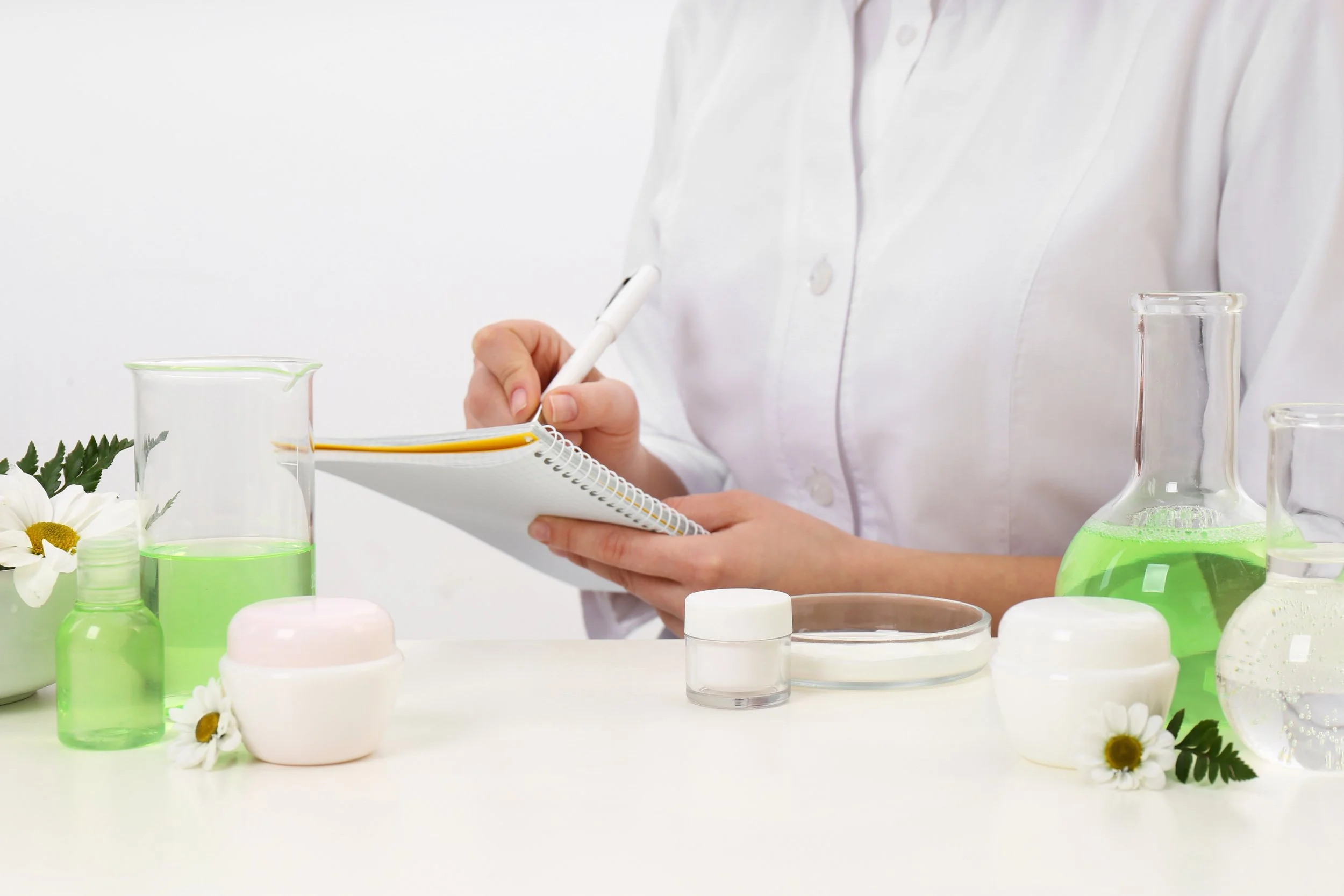 A person in a white shirt taking notes in a notebook surrounded by laboratory equipment containing green liquid, small bottles, jars, and flowers on a white surface.