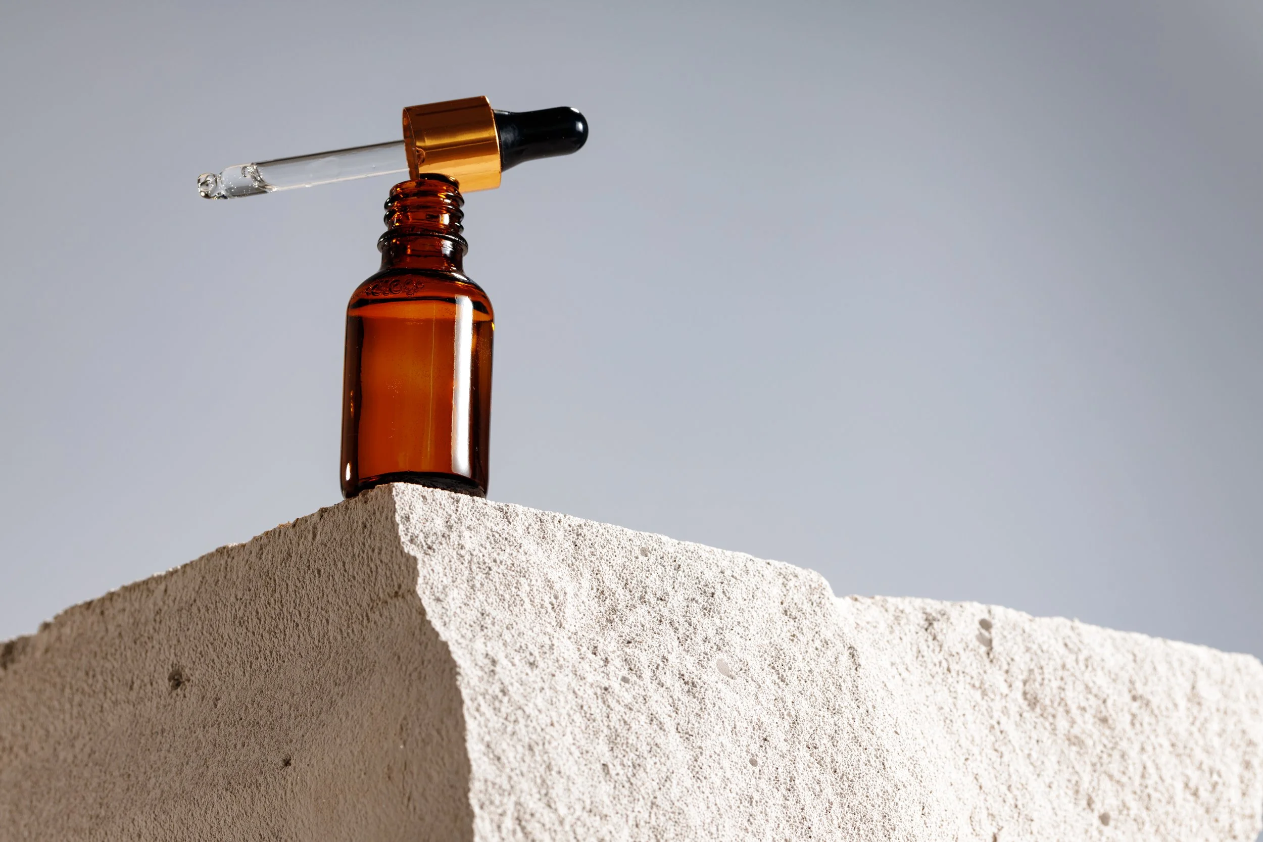 An amber glass dropper bottle with a black and gold cap, resting on a white textured surface with a dropper tube attached.
