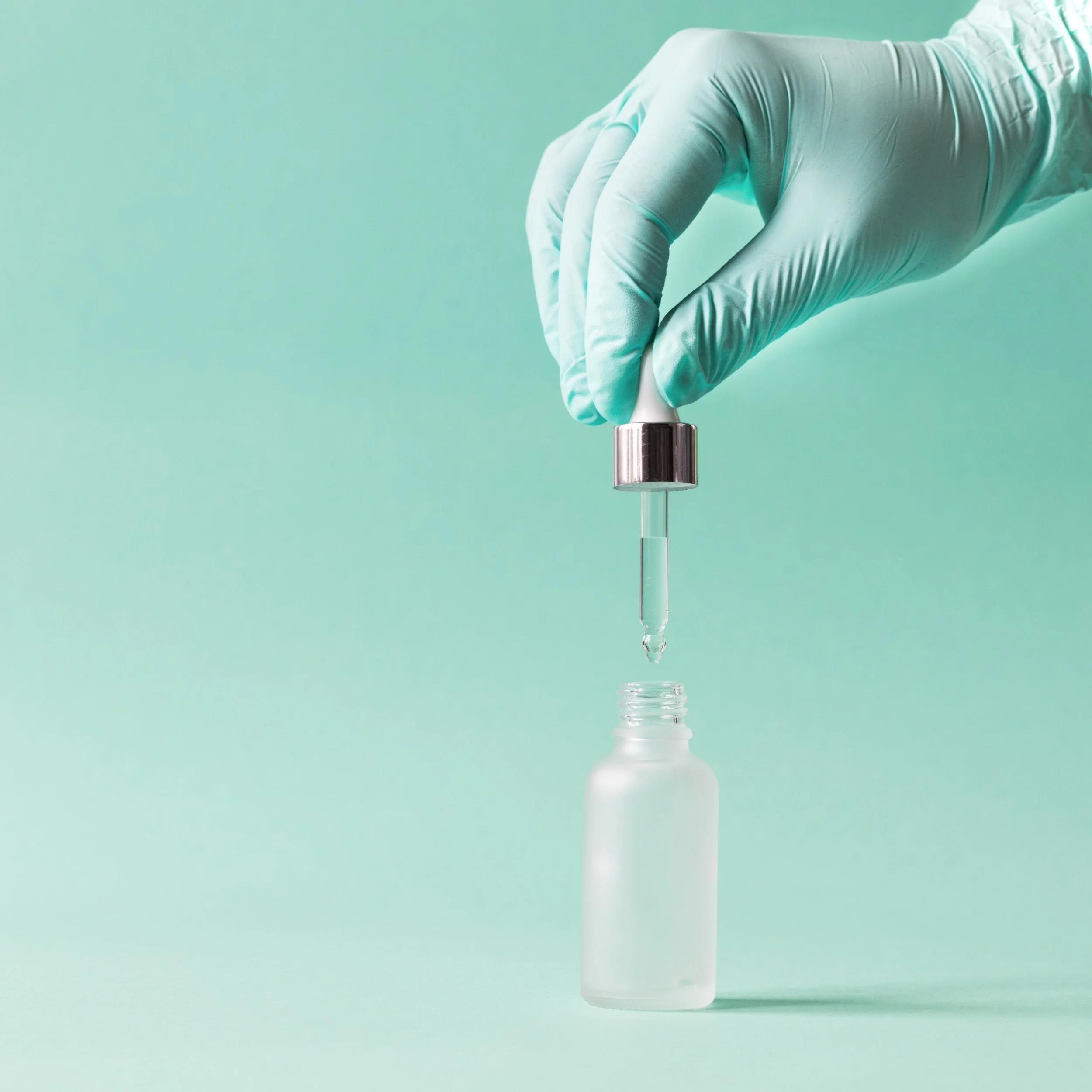 A hand in a medical glove holding a glass dropper with a droplet of liquid, positioned above a frosted glass bottle against a pastel green background.