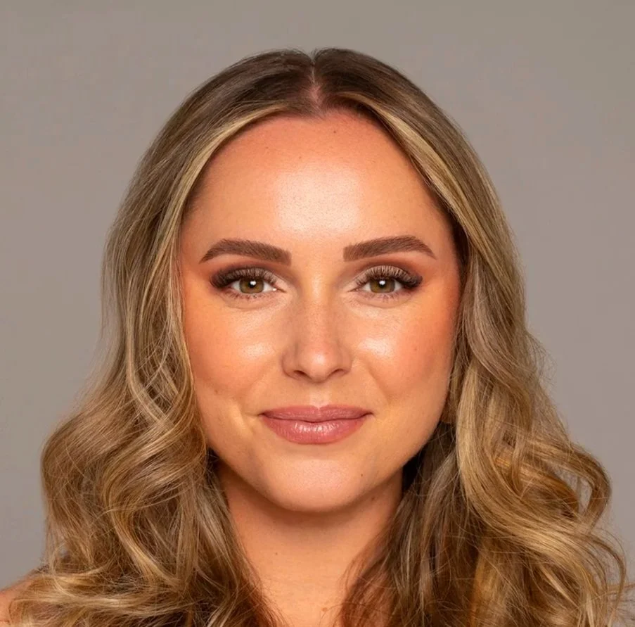 Close-up portrait of a woman with long, wavy blonde hair, wearing makeup, smiling softly, with a neutral gray background.