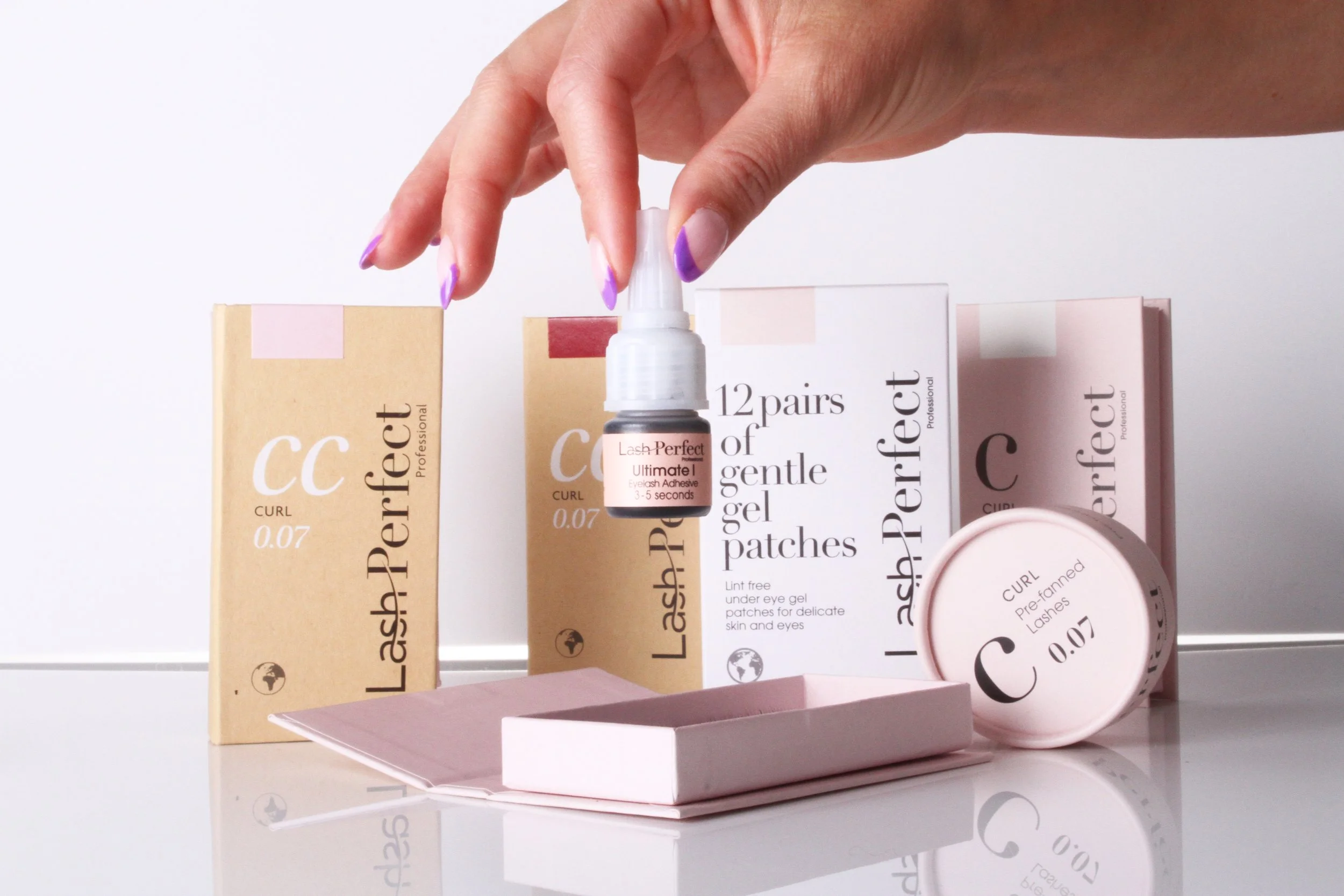 Collection of Lash Perfect eyelash extension products, including boxes of eyelash extension patches and a small bottle of eyelash adhesive, with a hand holding the adhesive bottle.
