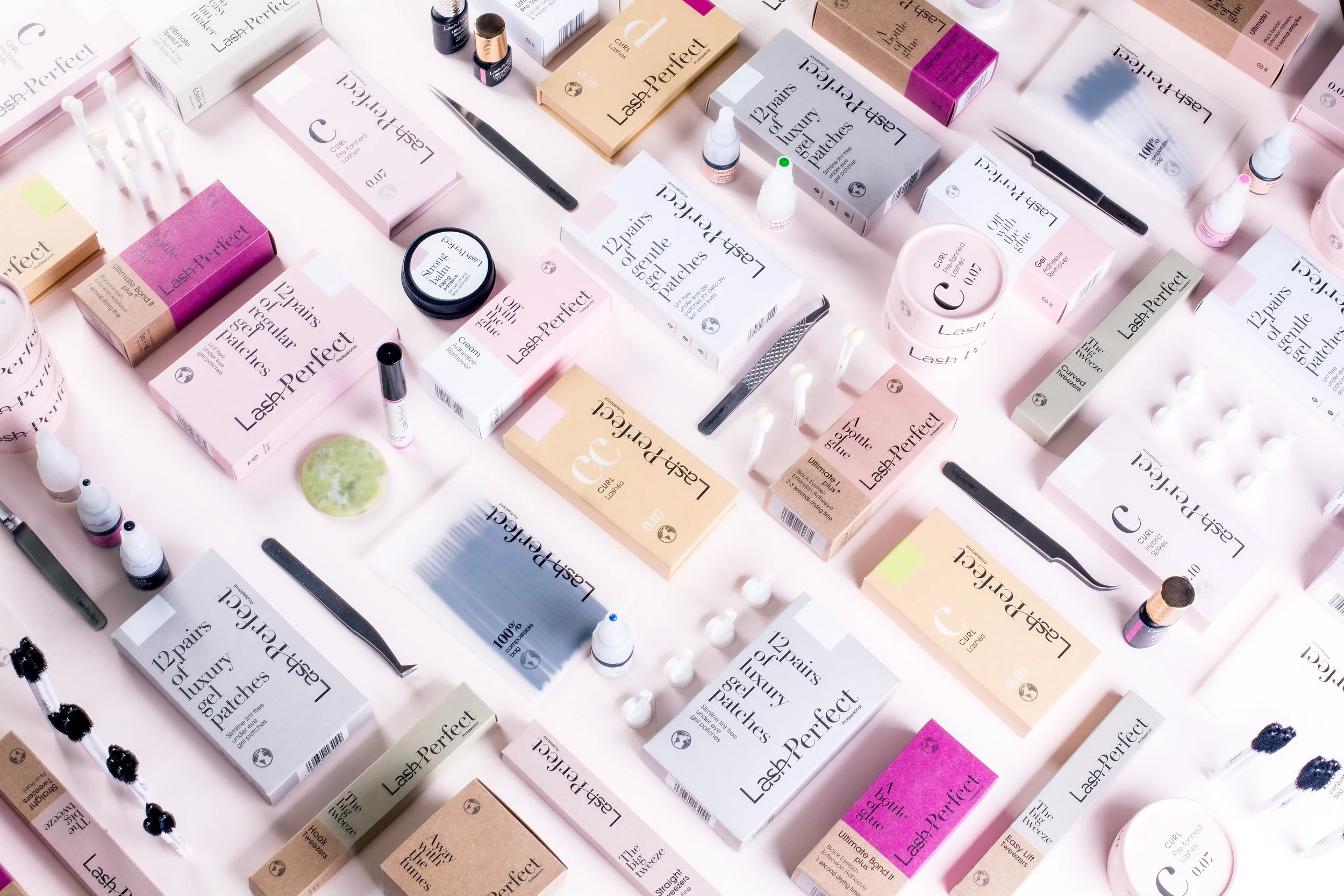 Assorted lash extension products and tools on a pink surface, including boxes of lash patches, glue, tweezers, and applicators.
