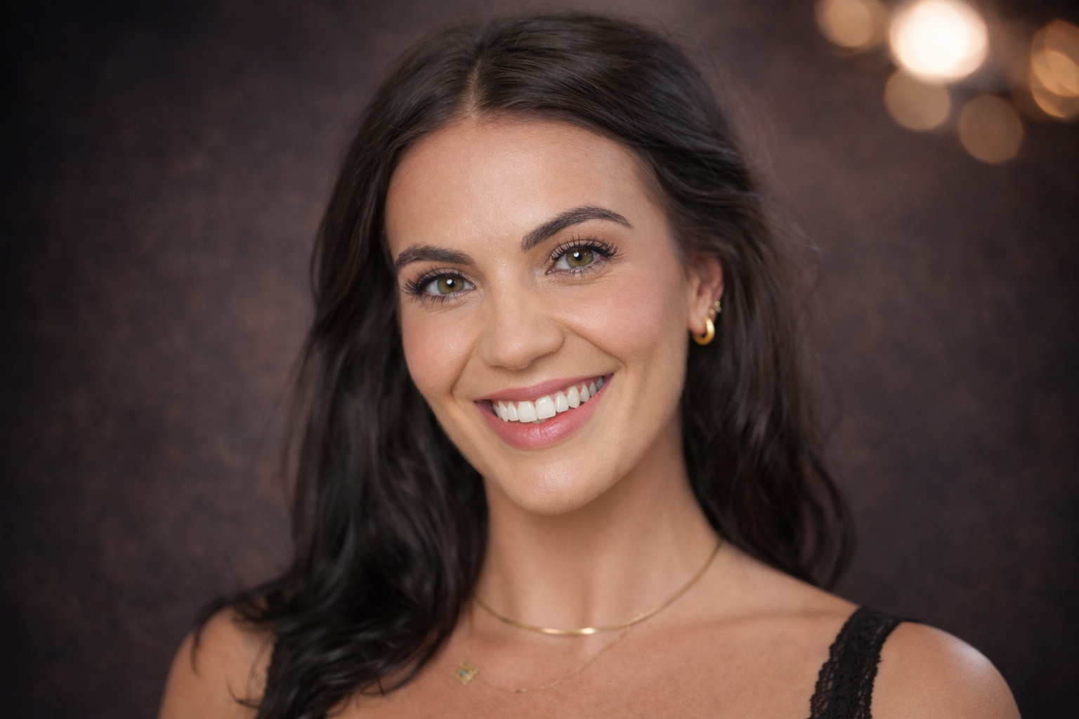 A woman with long dark hair, light skin, and hazel eyes smiling at the camera, wearing a black lace top, gold hoop earrings, and layered necklaces.
