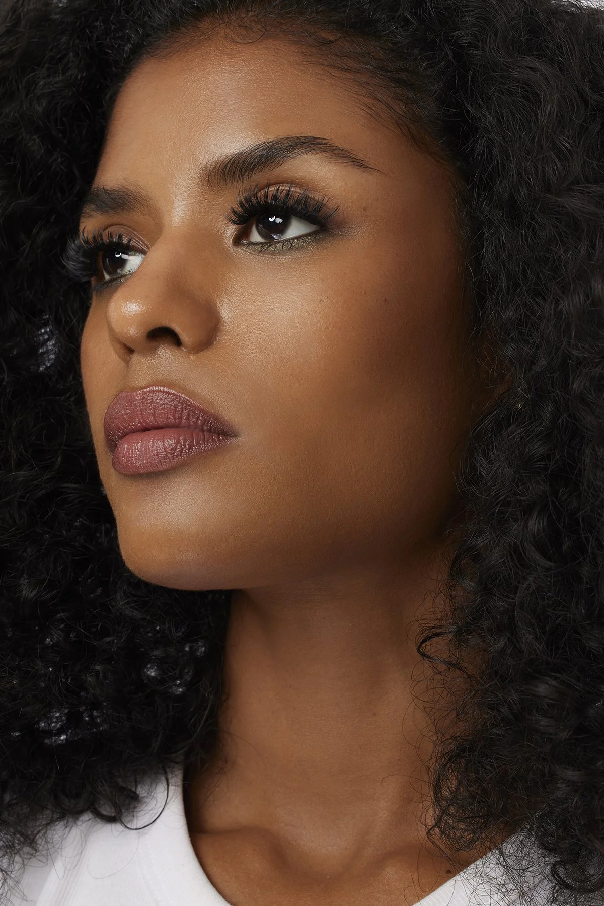 Close-up of a woman's face with curly black hair, wearing makeup including eyeliner, mascara, and lipstick, against a dark background.