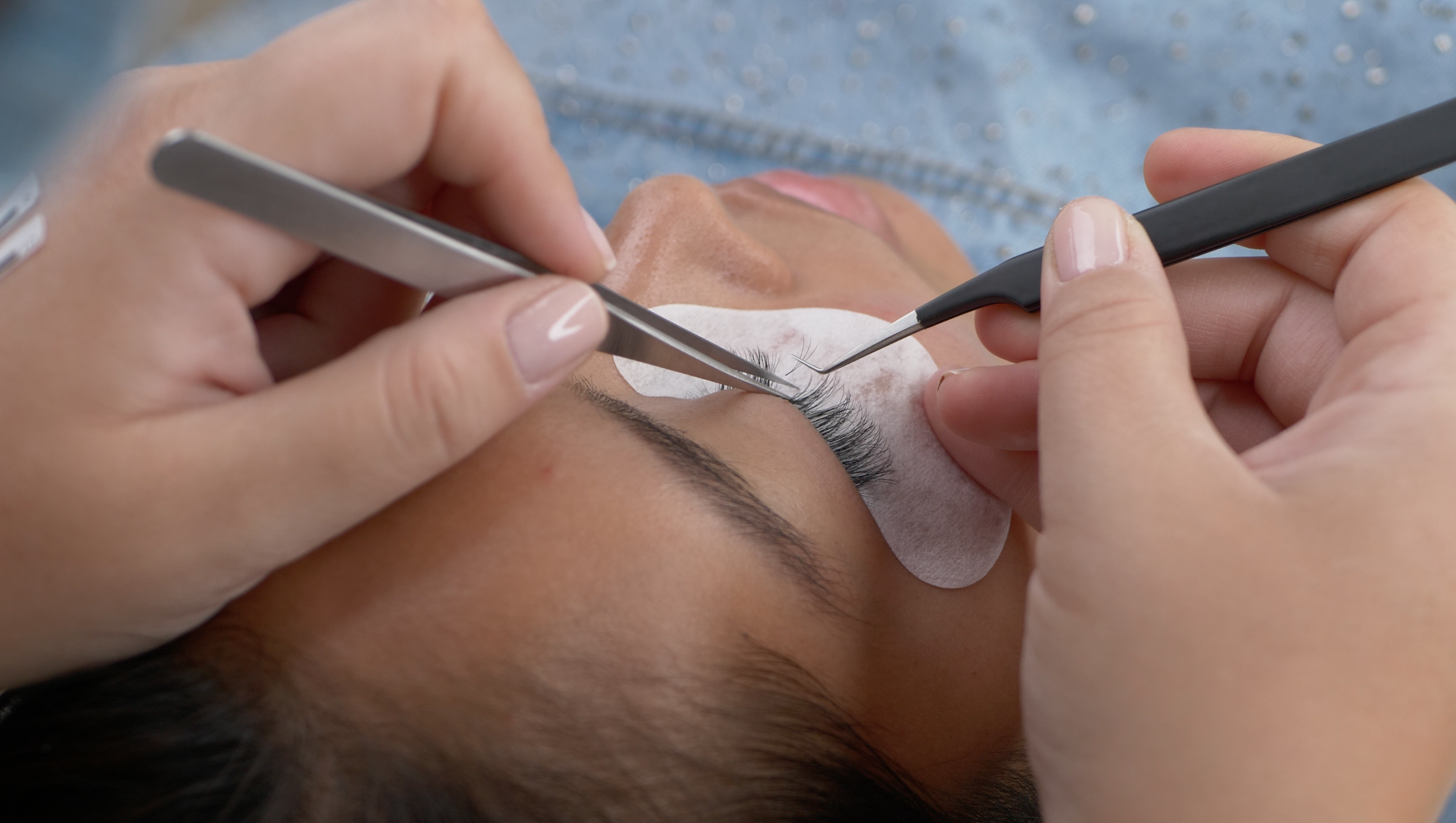 Close-up of a person receiving eyelash extension application with eyelash extension tools and protective pads over the lower eyelid.
